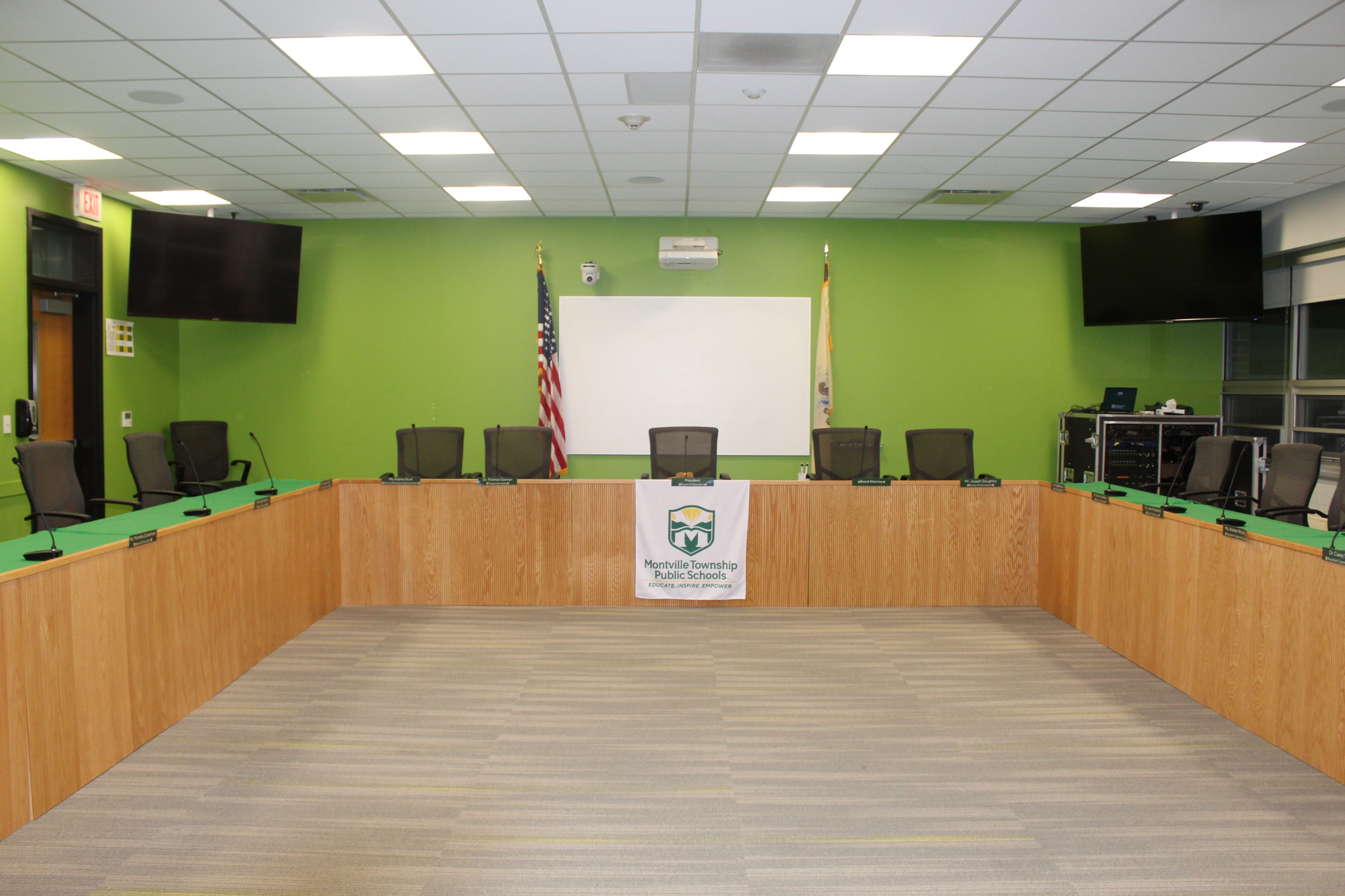 Spacious conference room with green walls, wooden tables, and black chairs arranged in a U-shape. Wall has U.S. flag and whiteboard. Bright atmosphere.