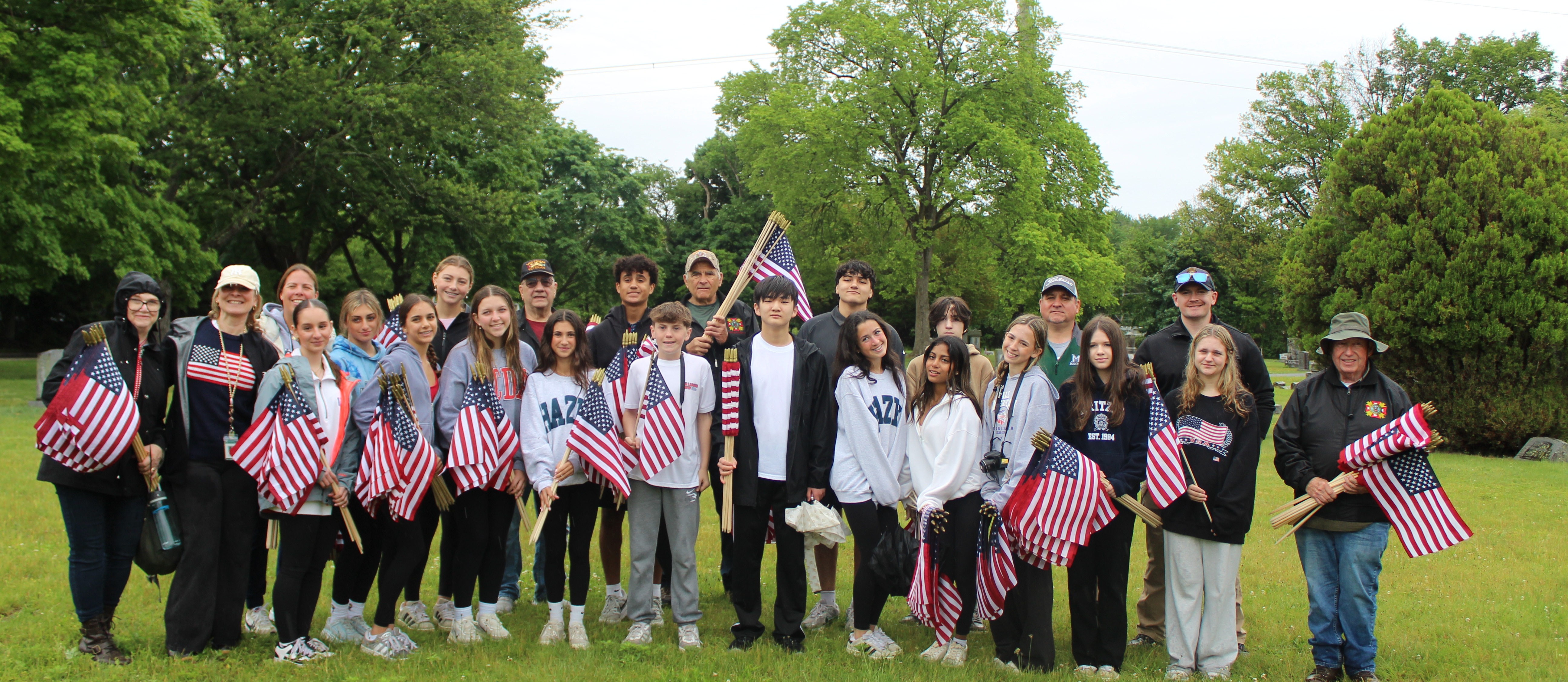 A group of people stand on a grassy field holding American flags. They are casually dressed, smiling, and surrounded by lush greenery and trees.