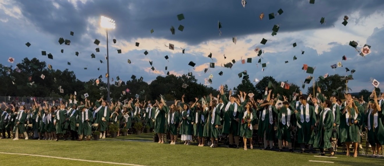 Graduates in green robes joyfully toss hats into the sky on a football field. A dramatic cloudy sky and bright stadium lights frame the celebratory scene.