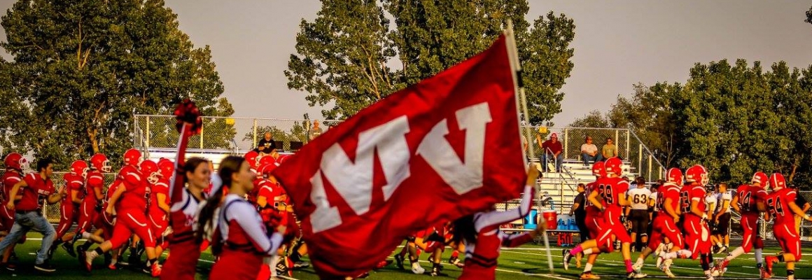 the football team and cheerleaders in red running across the field, with a red "MV" banner in the foreground