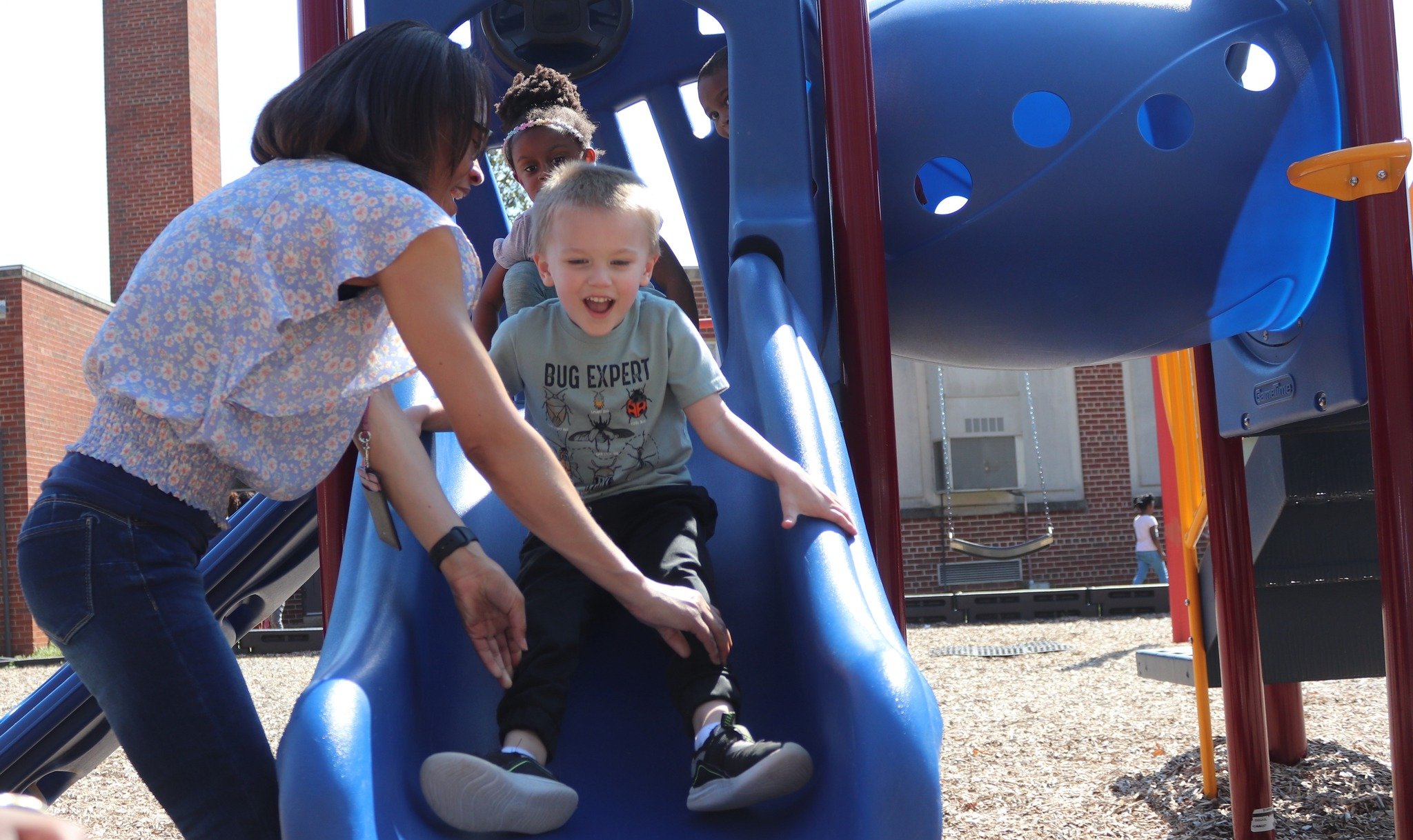 A preschool student smiles as he is helped down a slide by a teacher