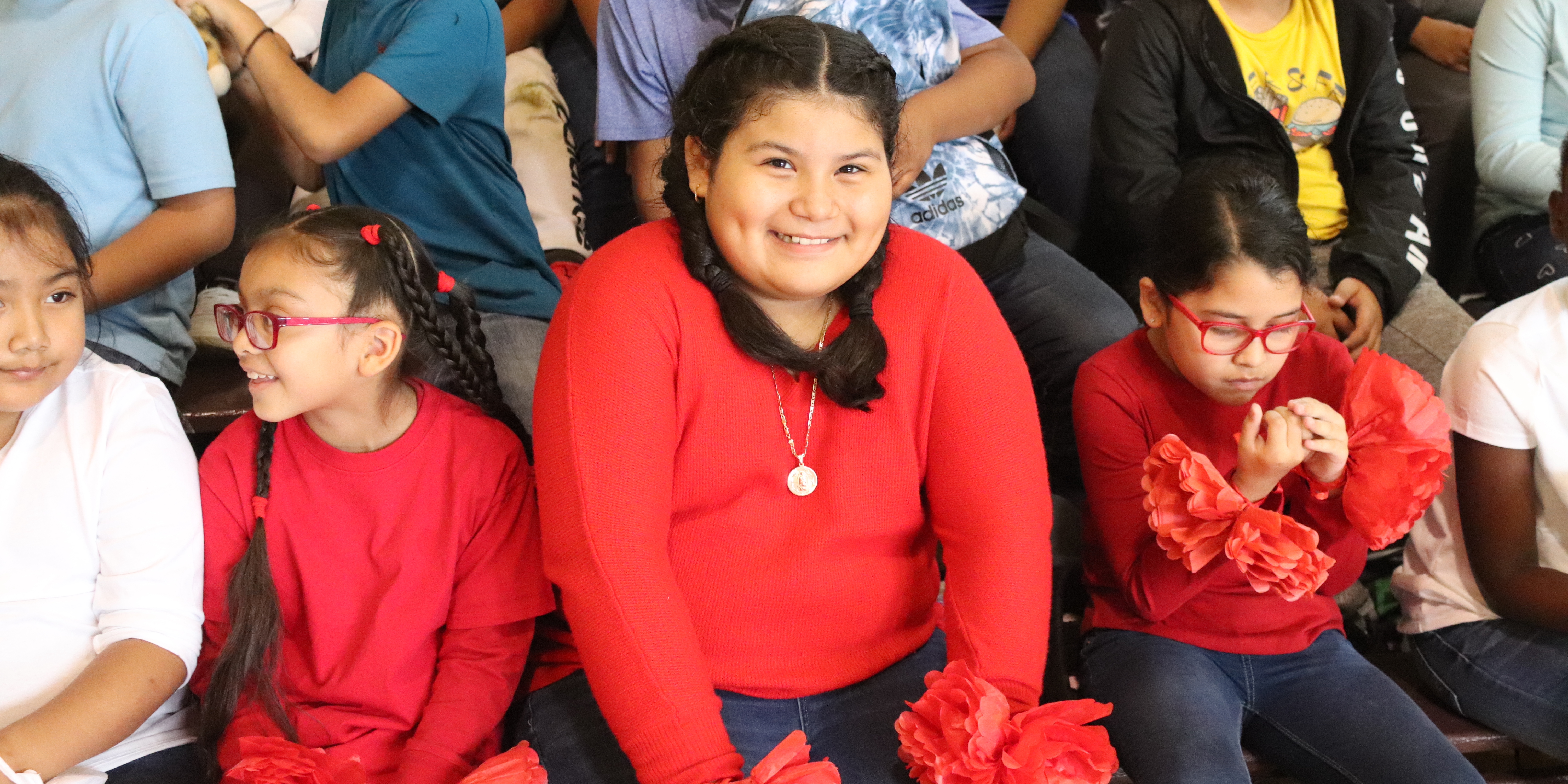 A crowd of elementary students sit on bleachers. One girl dressed in a bright red shirt smiles. 