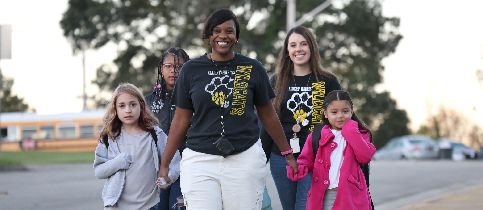 Teachers in Albert Harris t-shirts smile and hold the hands of students as they walk them from the bus into school