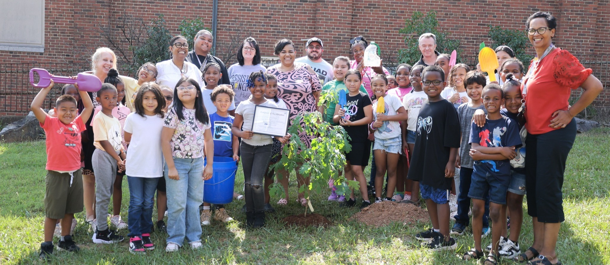 AHES students and teachers pose in a semi-circle around a newly-planted tree