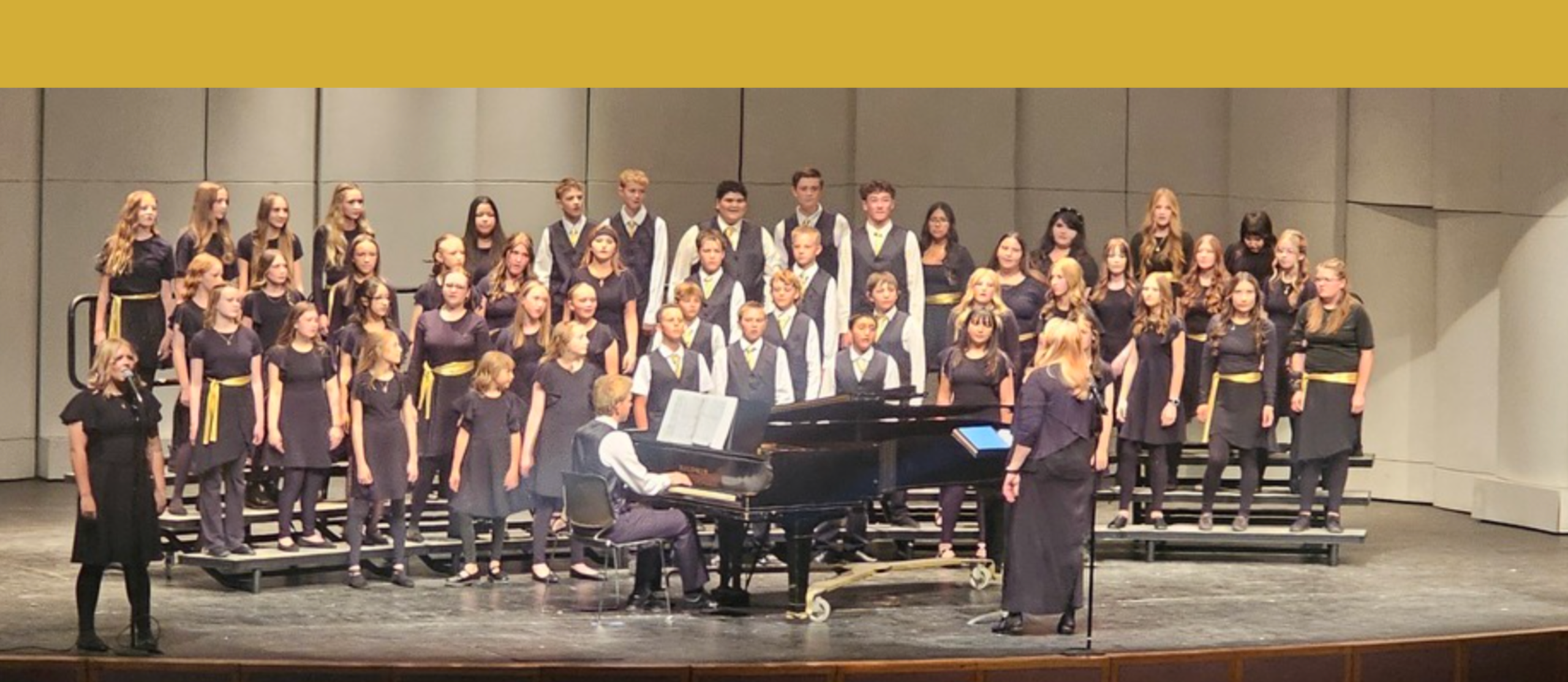 A choir of middle school kids on platforms singing with a grand piano in front of them with a student pianist and also a lead singer in the front to the right with a chorister / teacher slightly to the right of center.