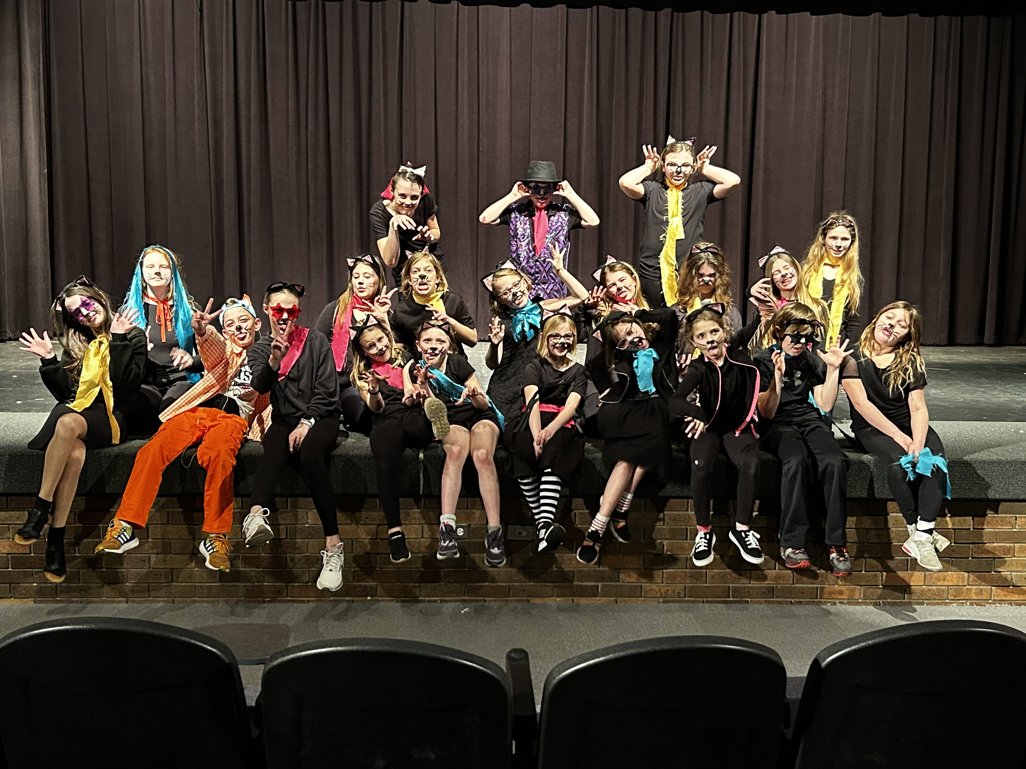 A large group of children in cat costumes and face paint pose playfully on a stage in front of a dark brown curtain, wearing various feline ears, tails, and colorful accessories.