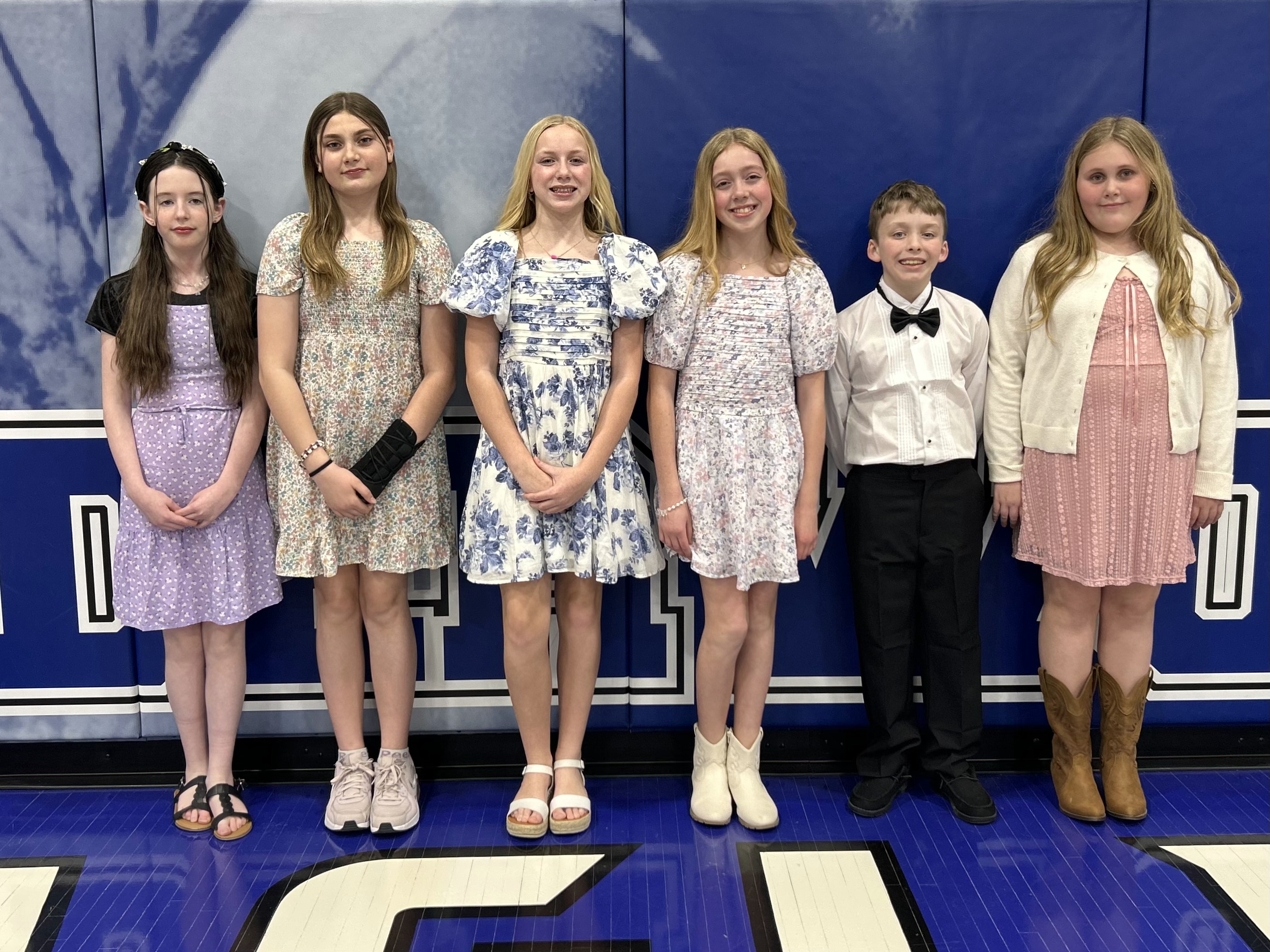 Six children dressed in formal attire, including floral dresses and a boy in a white shirt and bowtie, stand in a line against a blue and white athletic wall padding.