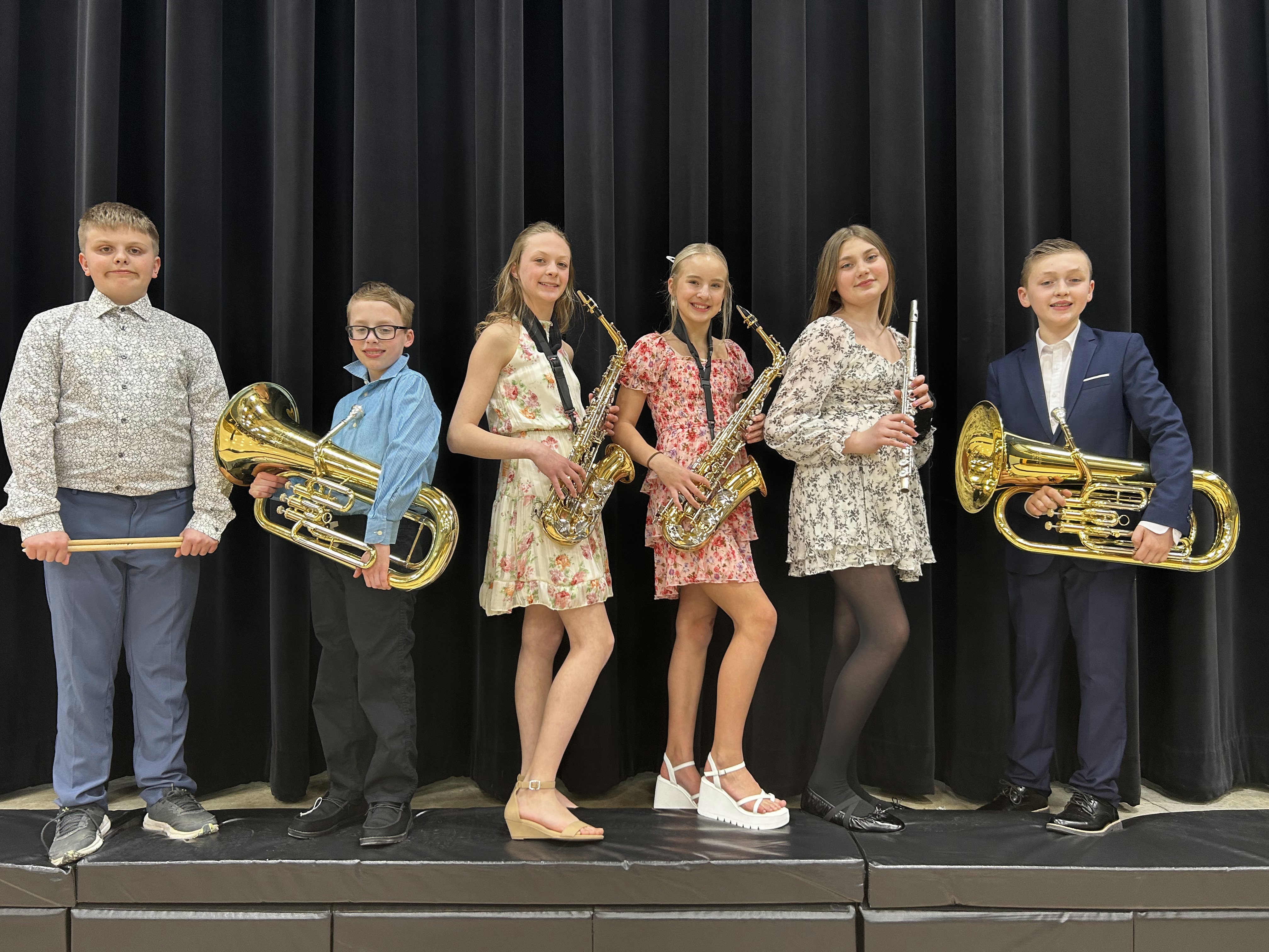 Six students in formal wear pose against a black curtain while holding musical instruments, including a drumstick, two euphoniums, two saxophones, and a flute.