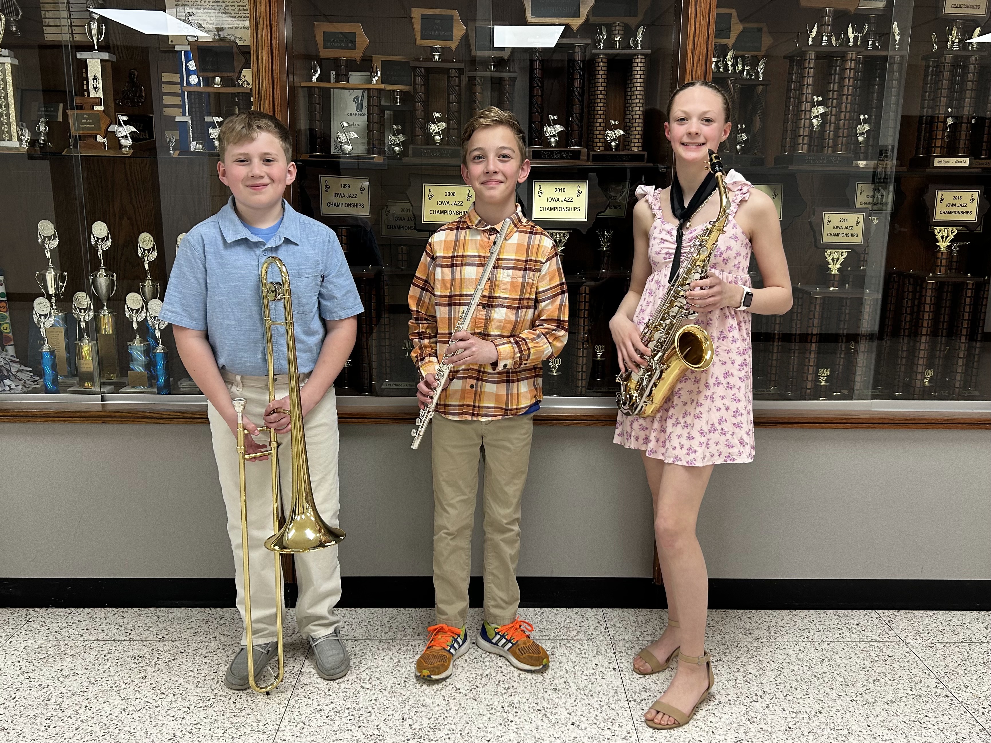 Three students pose with their musical instruments—a trombone, a flute, and a saxophone—in front of a large wooden trophy case filled with numerous awards.
