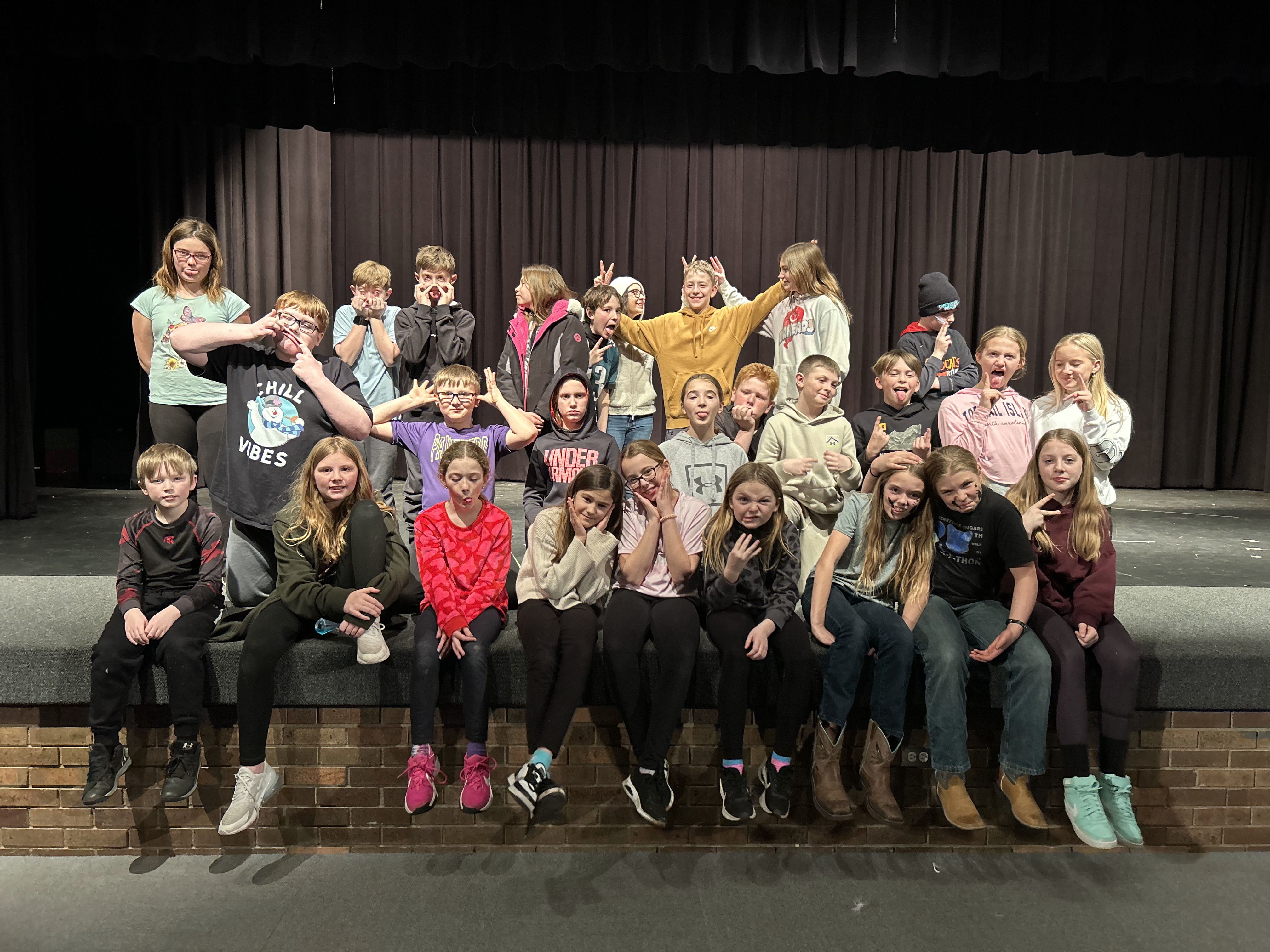 A group of students in casual clothes sit and stand on a stage in front of a dark curtain, making silly faces and hand gestures for a candid group photo.