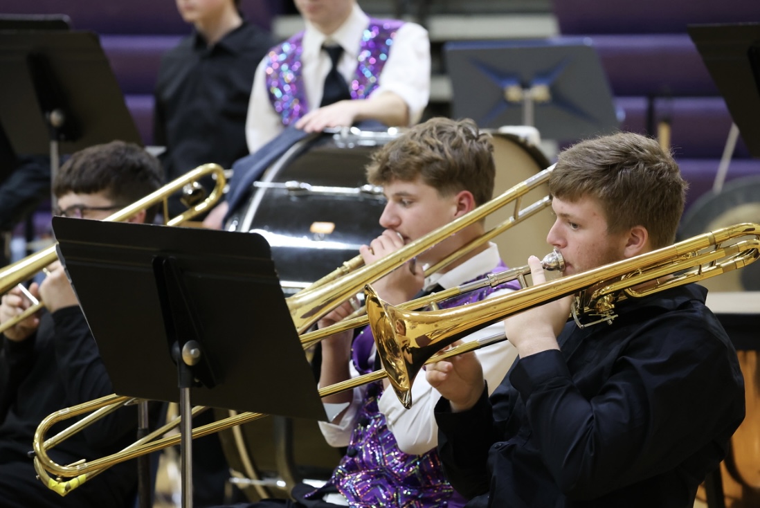 A group of trombone players perform in a row, with the center musician wearing a vibrant, sequined purple vest.