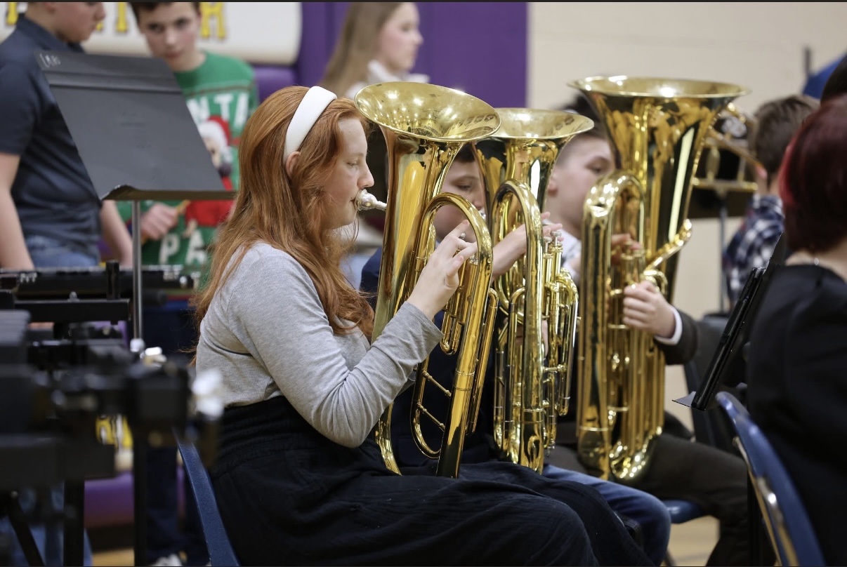 A close-up profile shows a young woman with long red hair and a white headband playing a large brass euphonium.