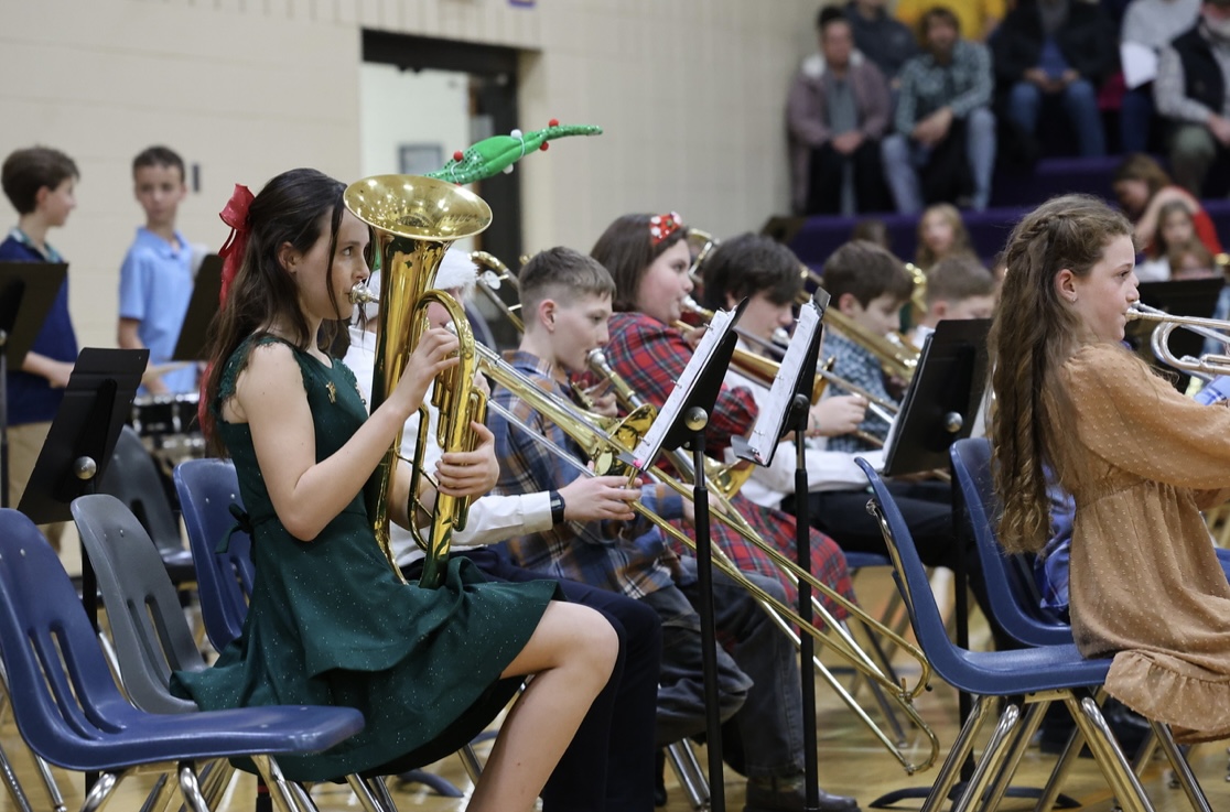 Student musicians in festive holiday outfits play brass instruments, featuring a girl in a dark green dress playing a euphonium.