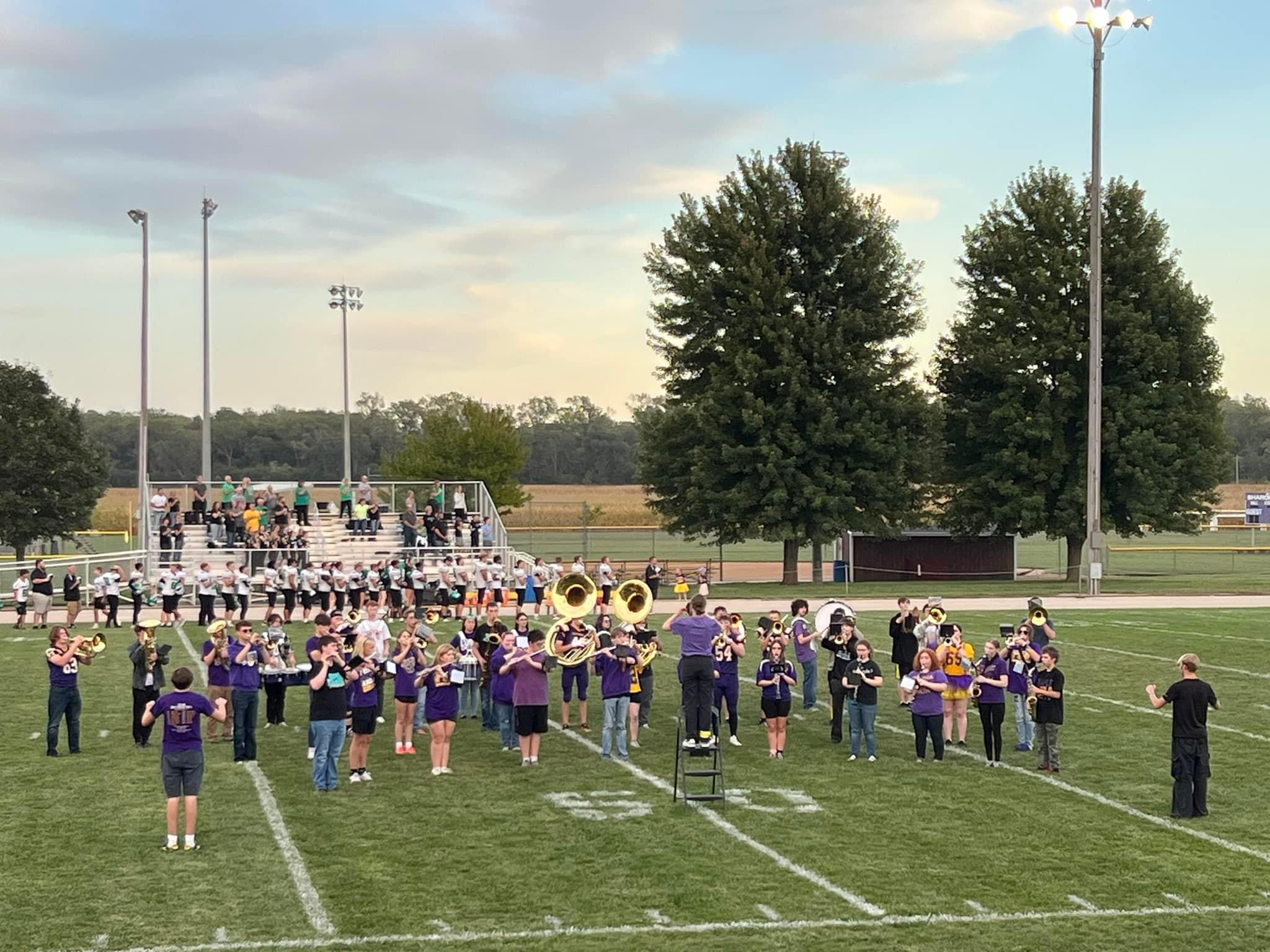 A marching band in purple shirts practices their formation on a grassy field at sunset, led by a conductor standing on a tall ladder.