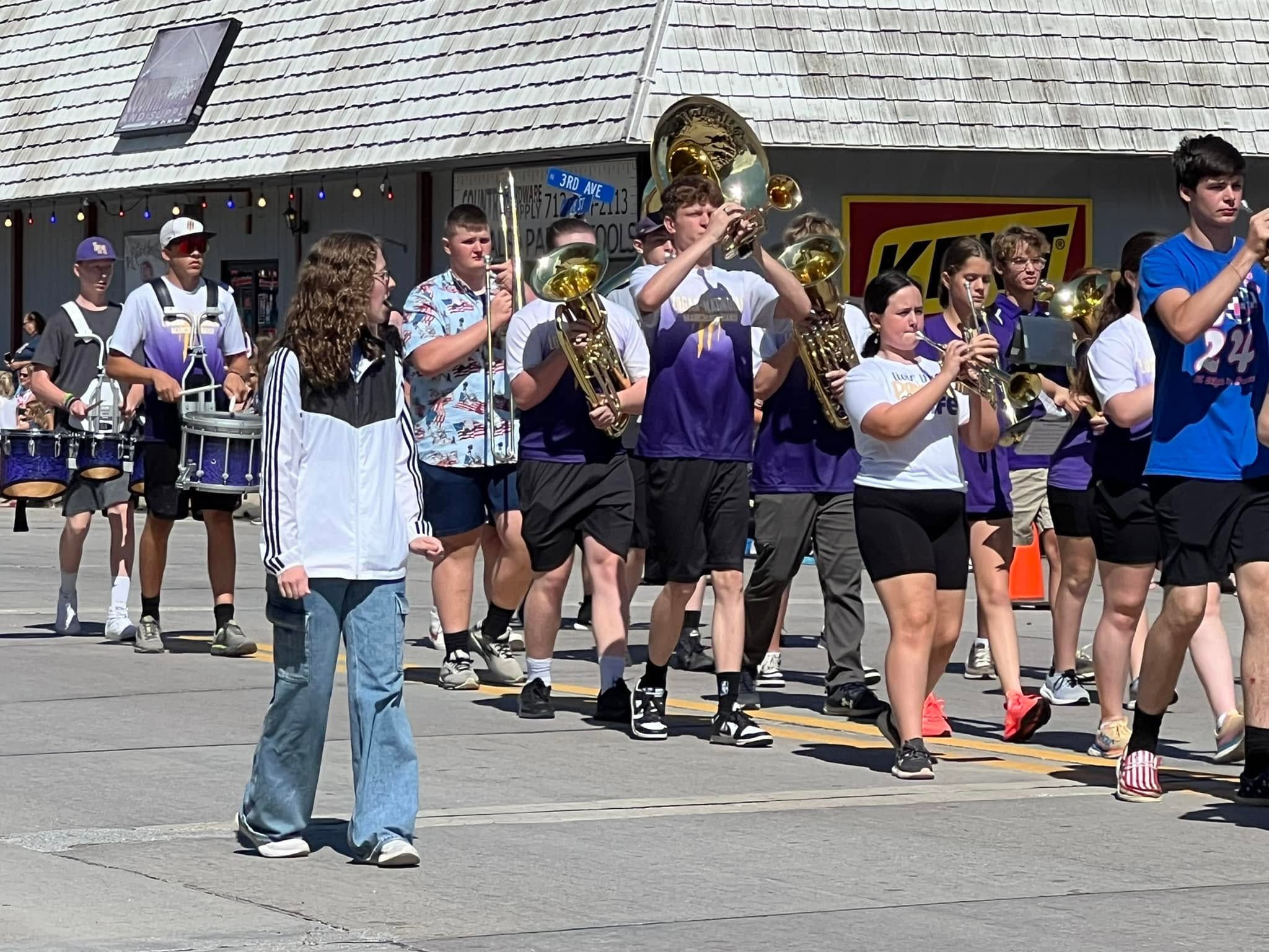 A marching band in casual purple and white gear performs with brass and percussion instruments while parading down a sunny street.