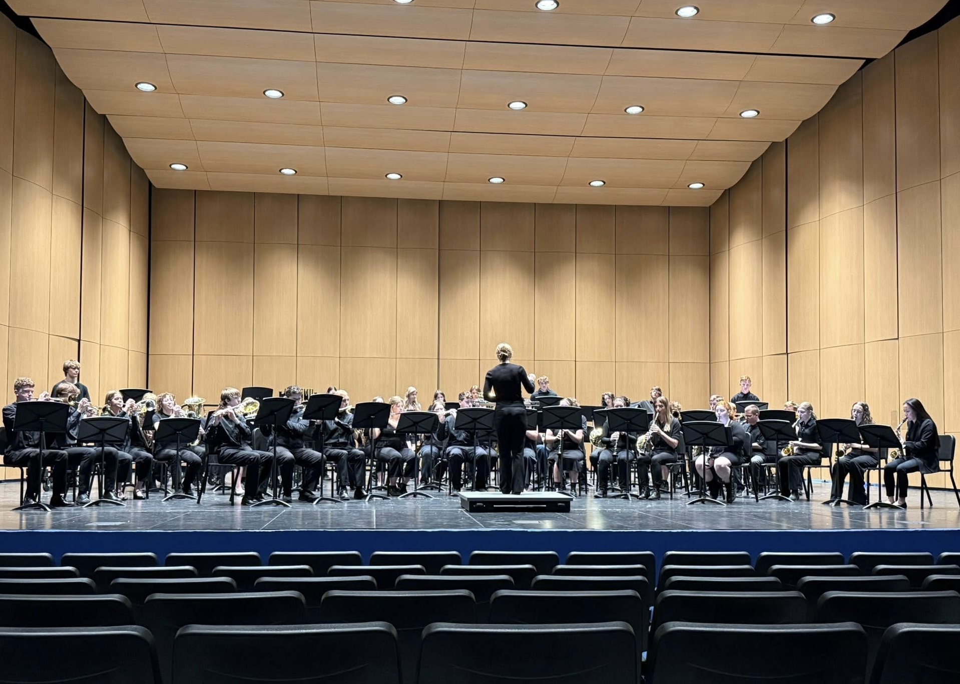 A large concert band dressed in formal black attire performs on a modern, wood-paneled stage under bright overhead lights.