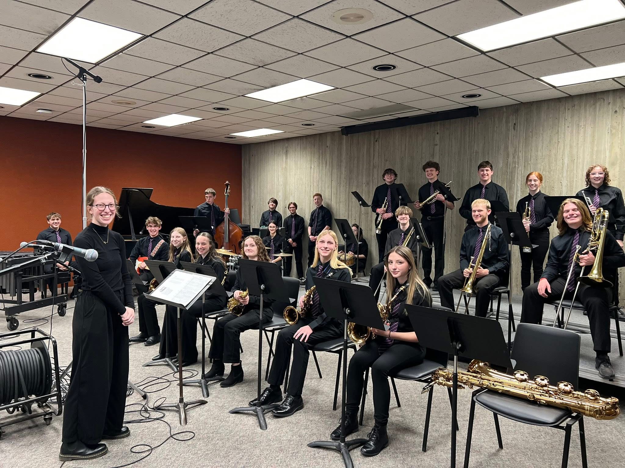 A jazz band dressed in black with purple ties poses for a group photo in a rehearsal studio alongside their instruments and conductor.