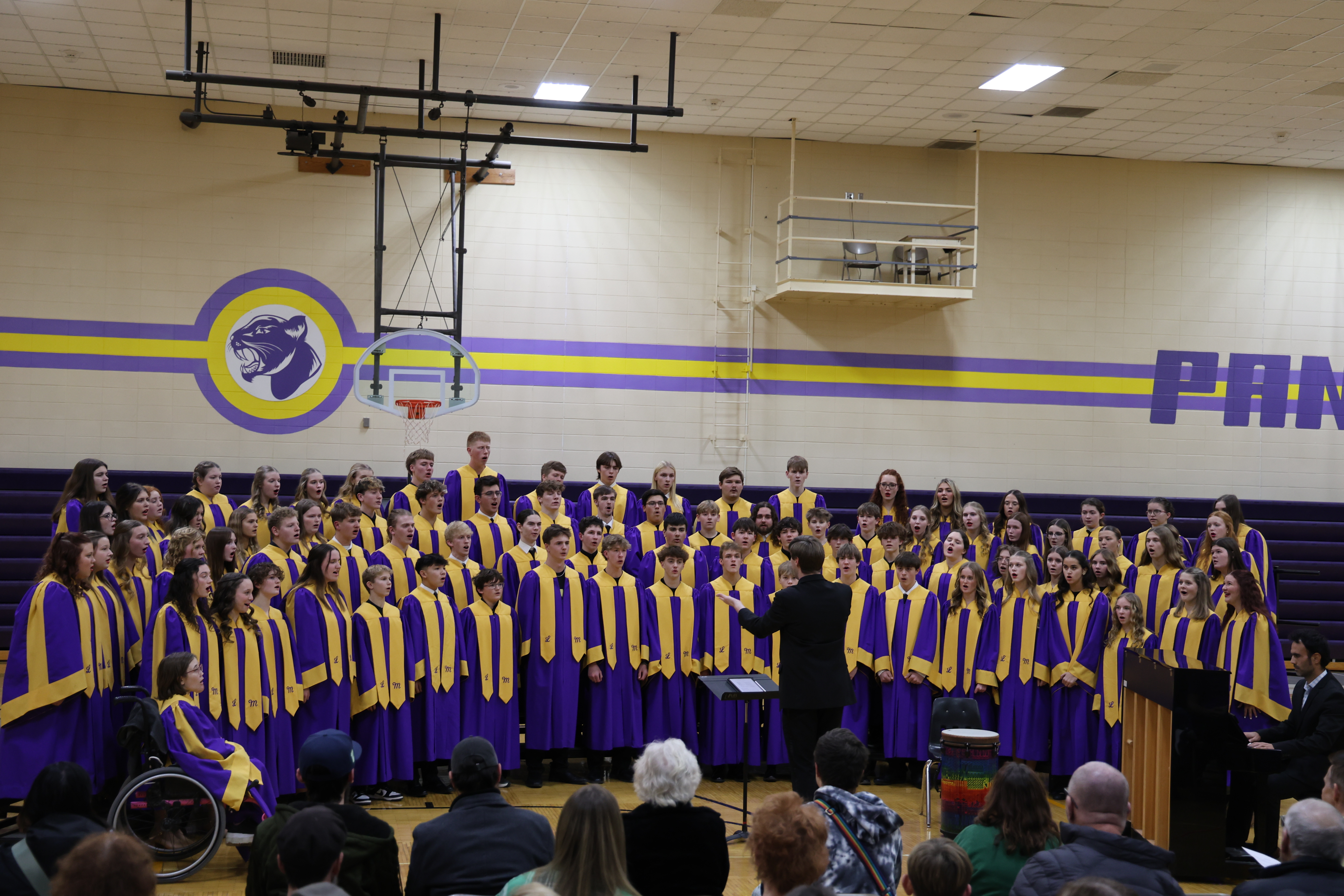 A full high school choir in purple and yellow robes performs on risers in a gymnasium, led by a conductor in front of a seated audience.