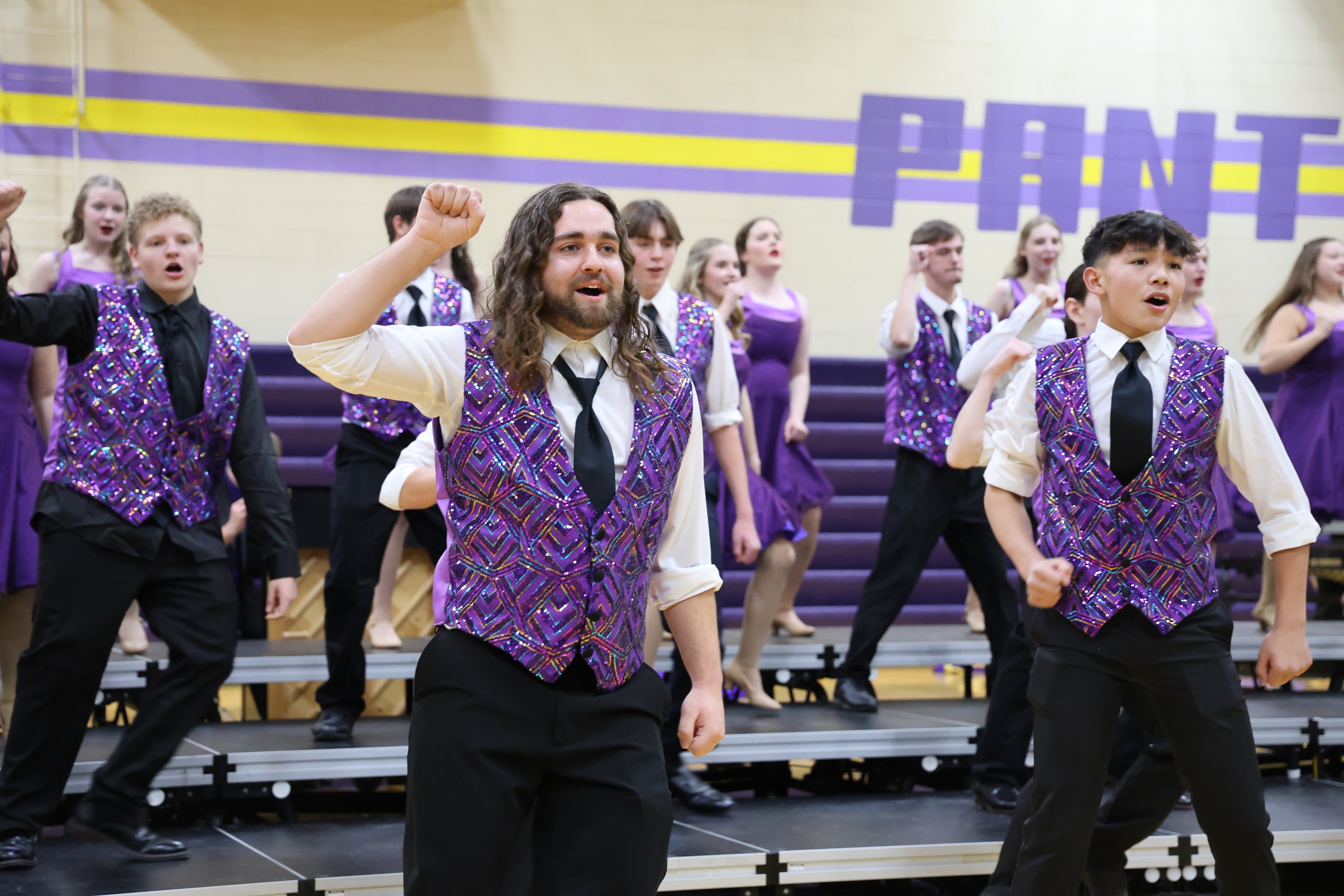 A high school show choir performer with long brown hair sings enthusiastically with a raised fist while wearing a sequined purple vest and black tie.