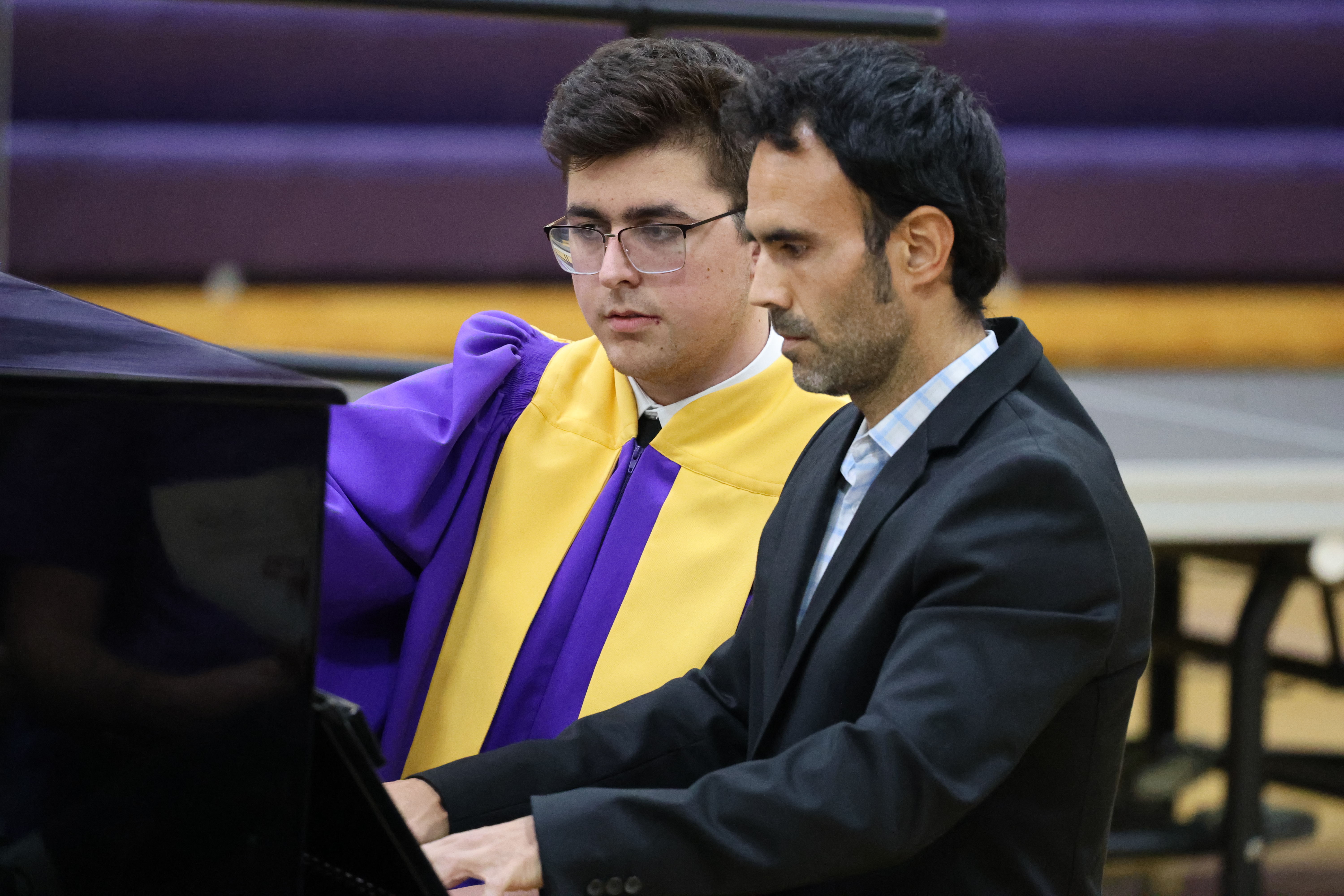 A choir student in a purple and yellow robe and a man in a black suit sit together at a black piano, looking intently at the keys.