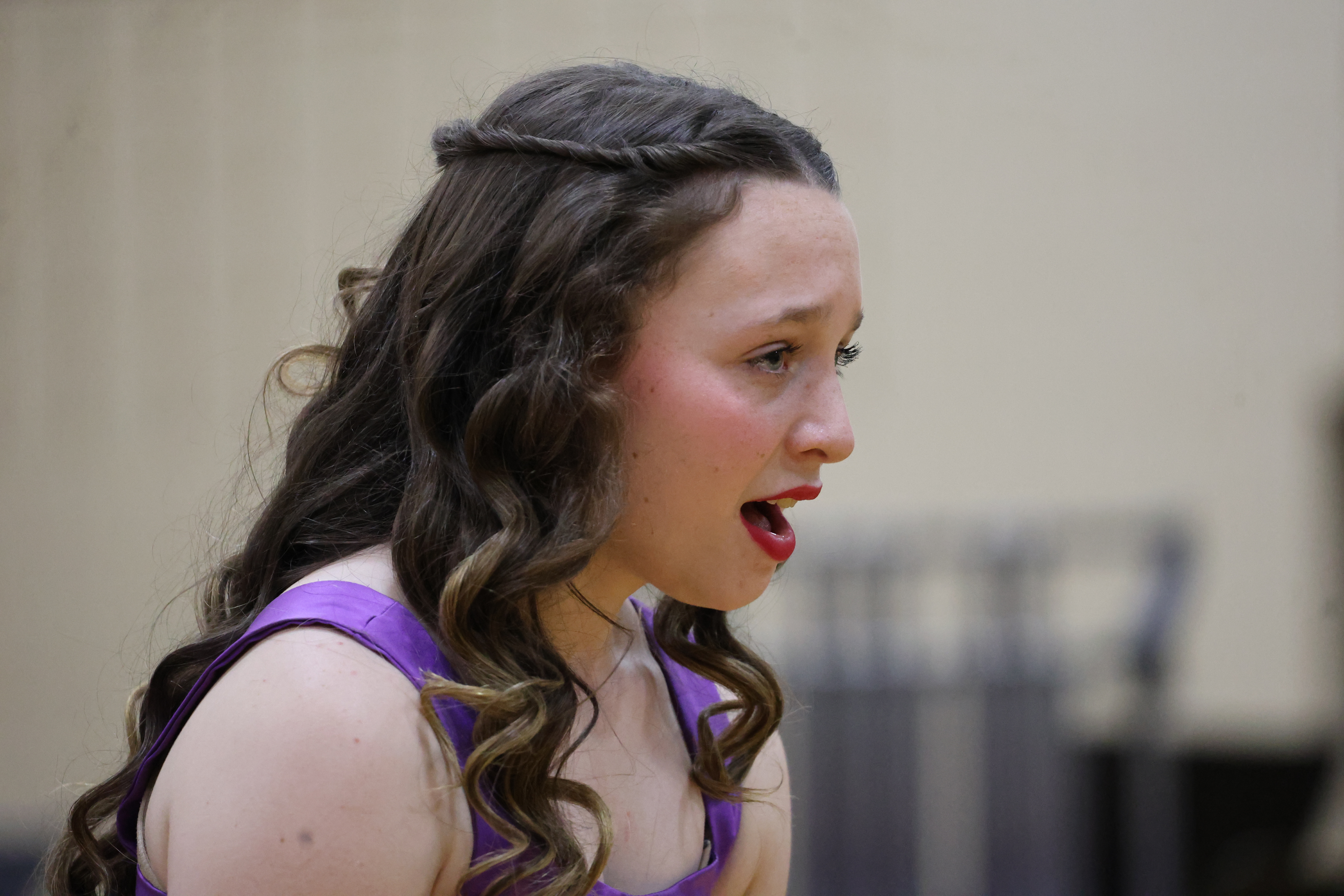 A close-up profile shot captures a young woman with long, curled brown hair and red lipstick singing with her mouth open during a performance.