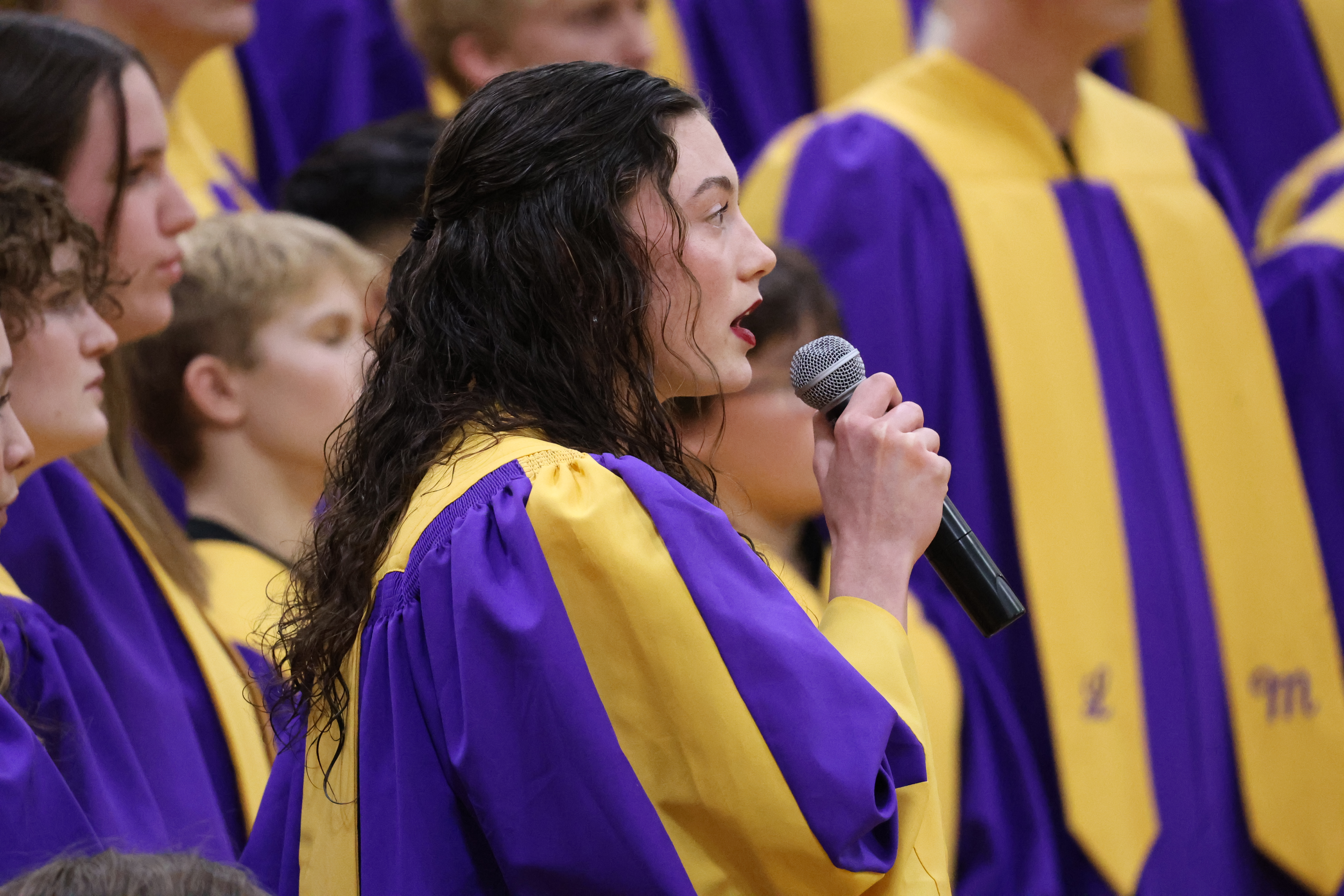 A young woman with long, dark wavy hair sings into a handheld microphone while wearing a purple choir robe with a yellow stole.