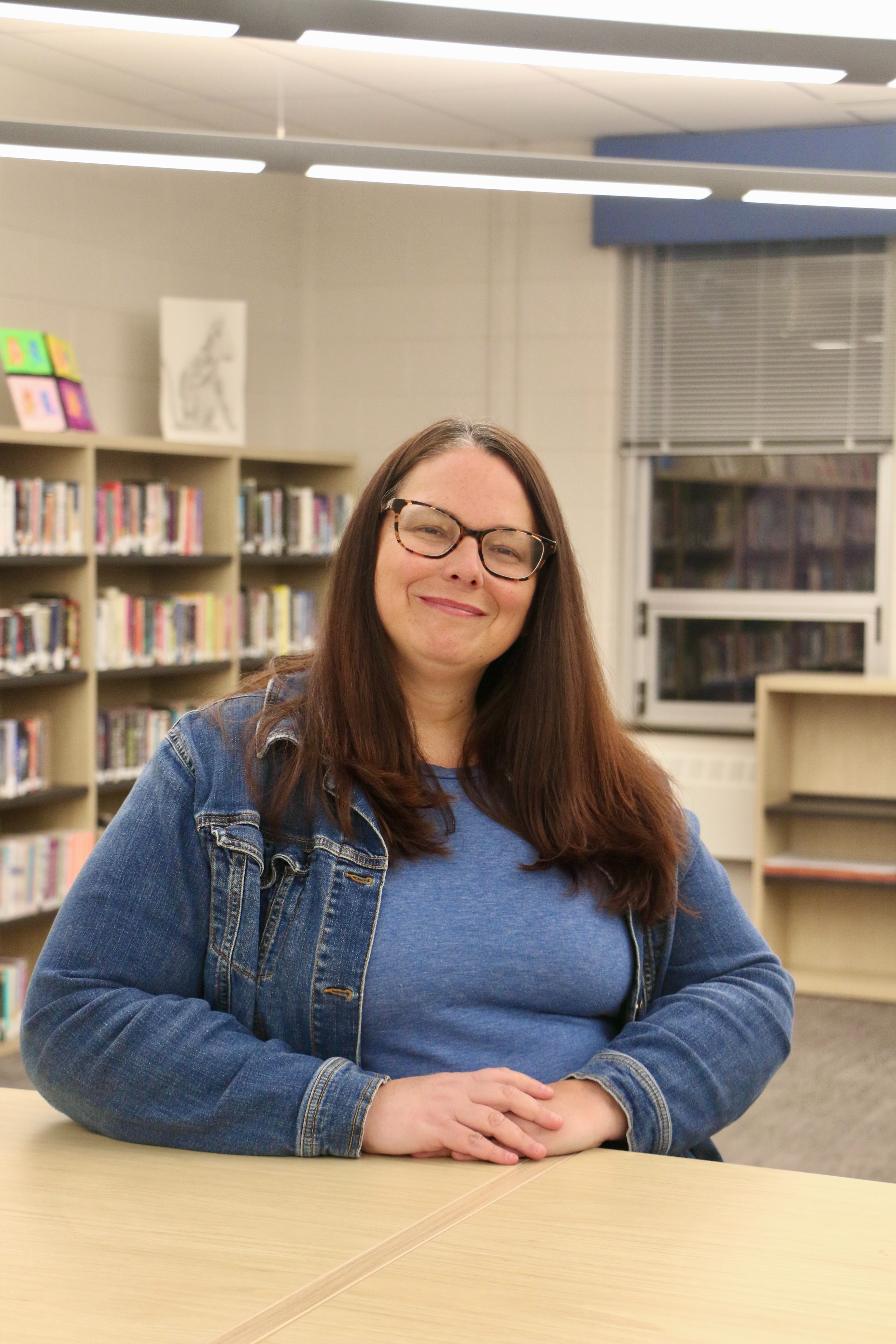 A woman with glasses smiles in a library with bookshelves and windows in the background.