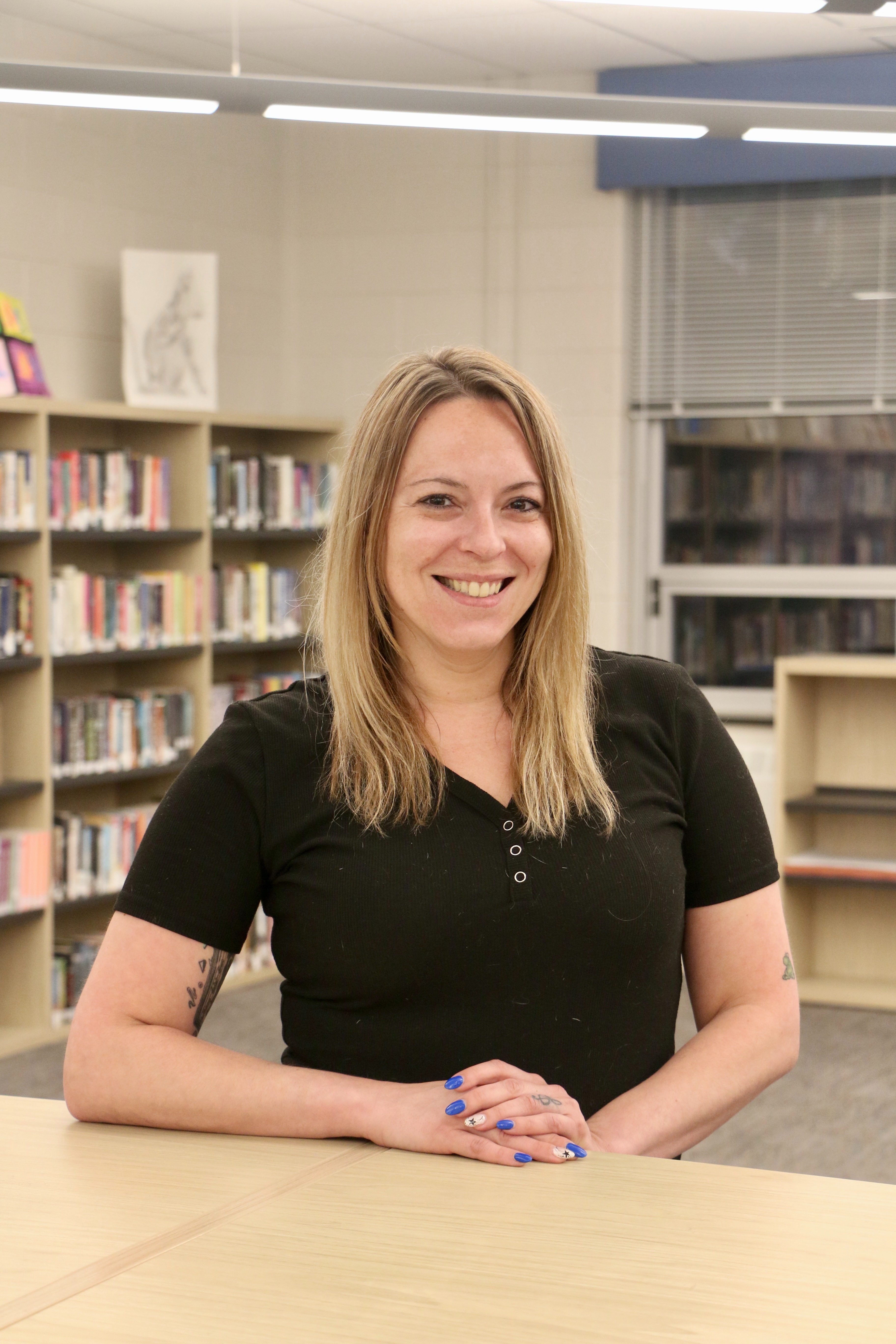 A woman with blonde hair smiles in a room with shelves filled with books and fluorescent lights.