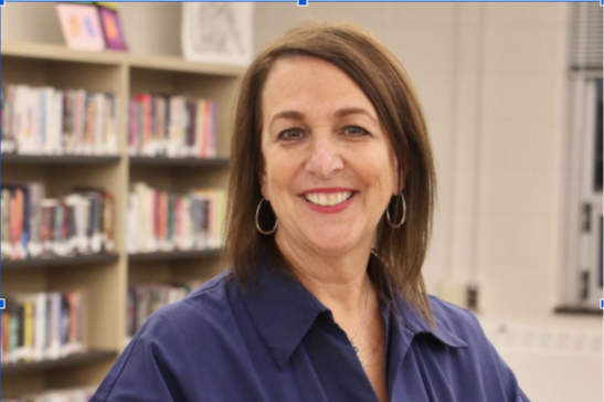 Woman in a blue shirt smiles at the camera in a room with bookshelves in the background.