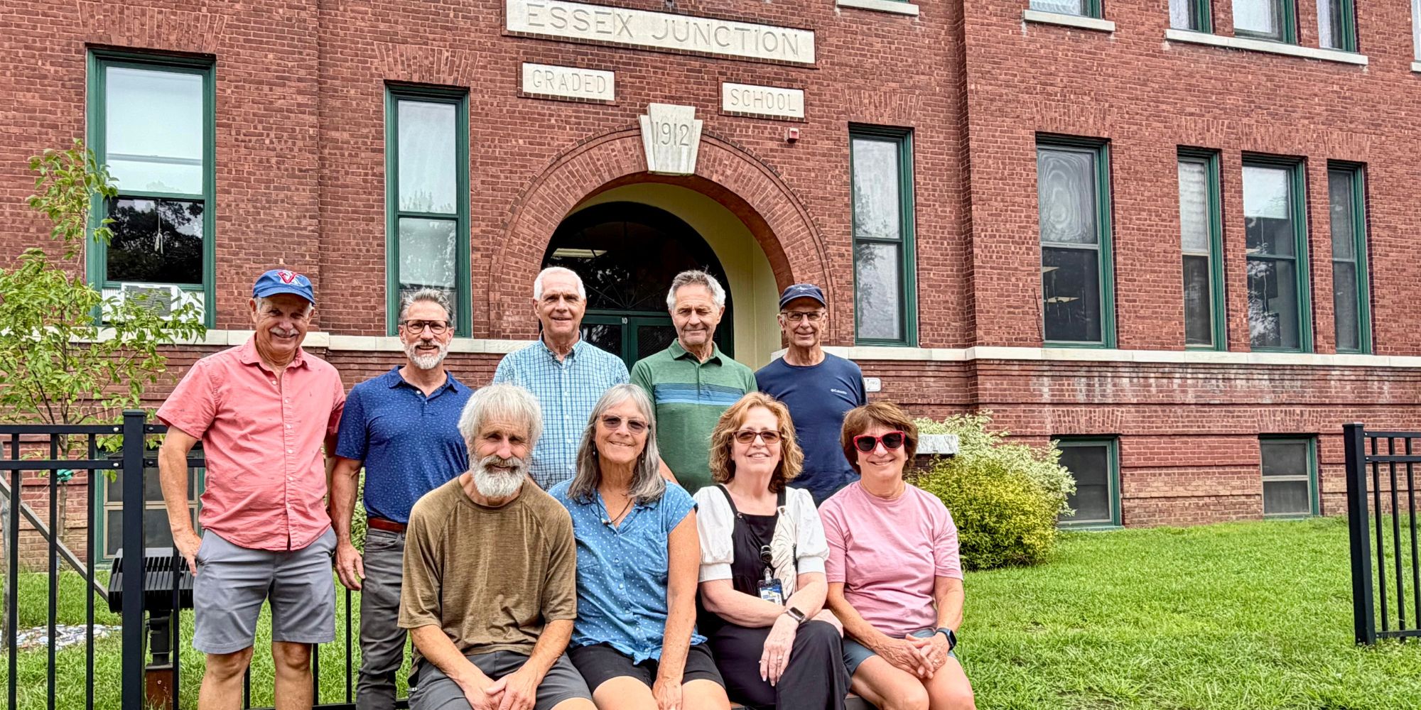 Group of people standing and sitting outside a brick building with arched doorway. Some wear hats, glasses, and sneakers.