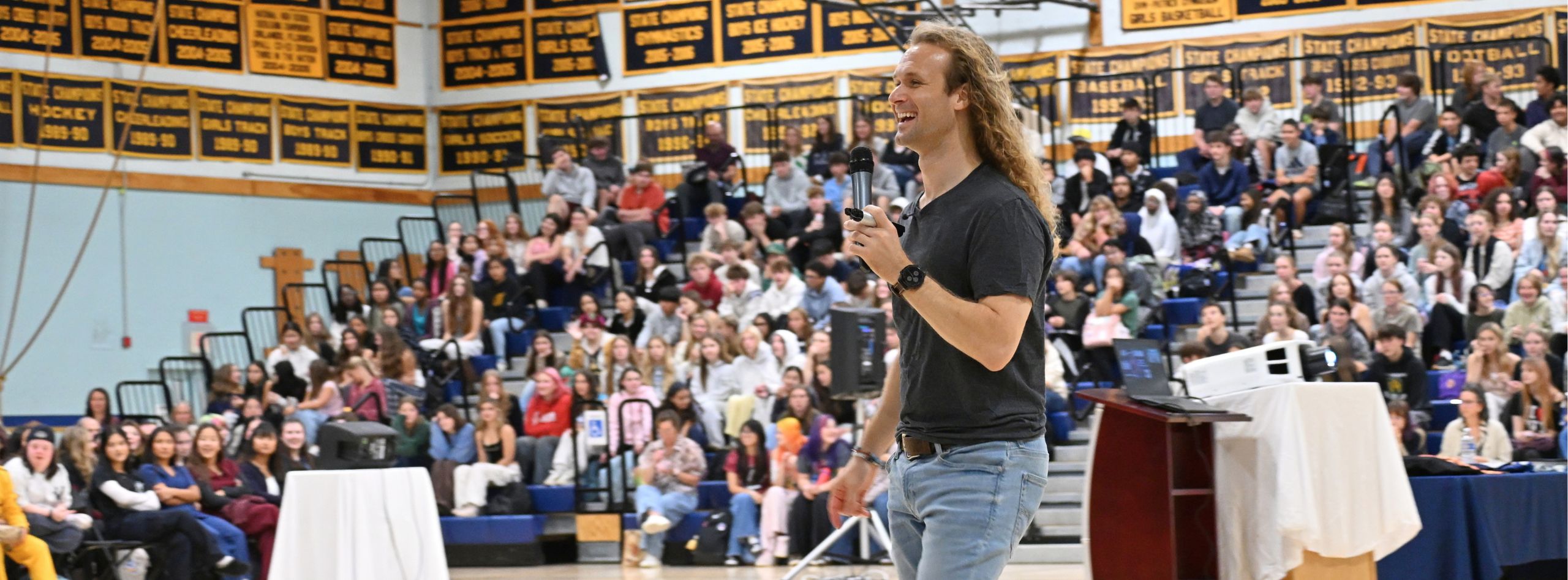 Man with long hair speaking into a microphone at a podium. A crowd of people are seated in the background.