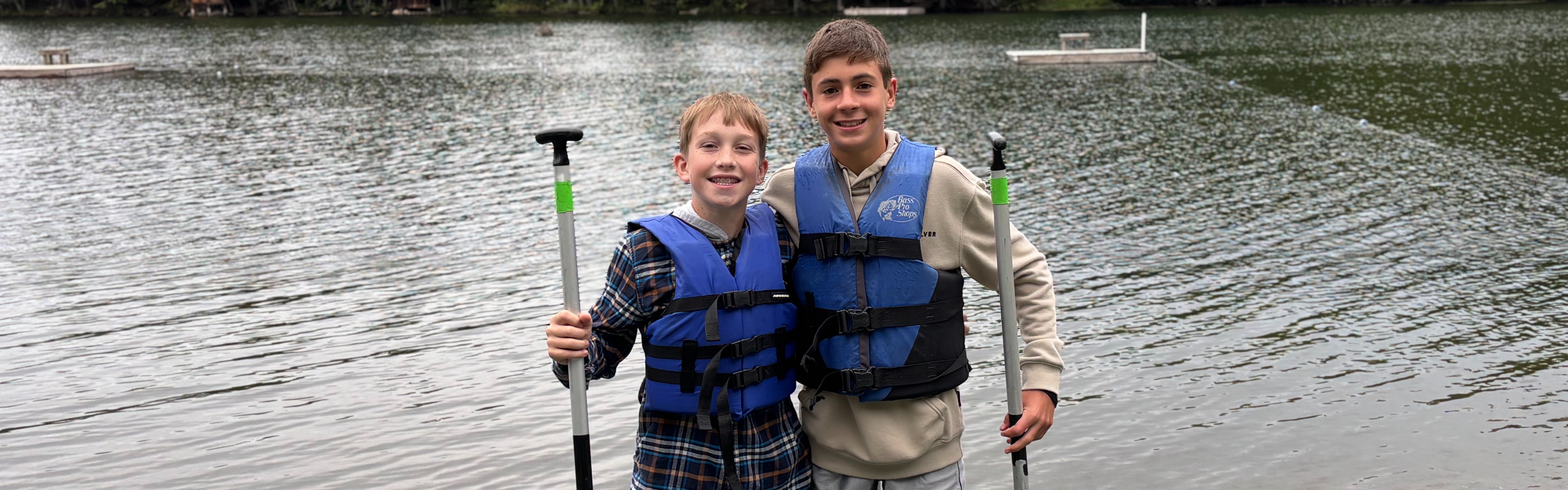 Two 8th grade boys wear blue life jackets and are holding paddles while they smile in front of Hosmer Pond