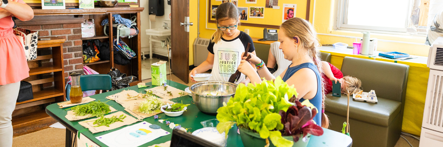 Two young children standing at a desk preparing a salad