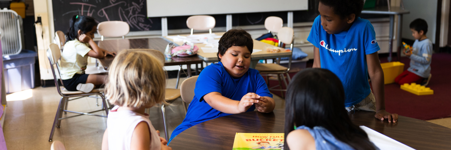 A group of young children in a classroom are engaged in different activities. In the foreground, four children sit around a table, with one boy in a blue shirt animatedly talking and gesturing with his hands while the others listen. A book titled How Full Is Your Bucket? is on the table. In the background, another child is sitting at a desk working, while a boy in a blue shirt stands nearby, watching the group at the table. Another child plays with large plastic building blocks on the floor. The classroom has natural light, a chalkboard with drawings, and various educational materials scattered around.