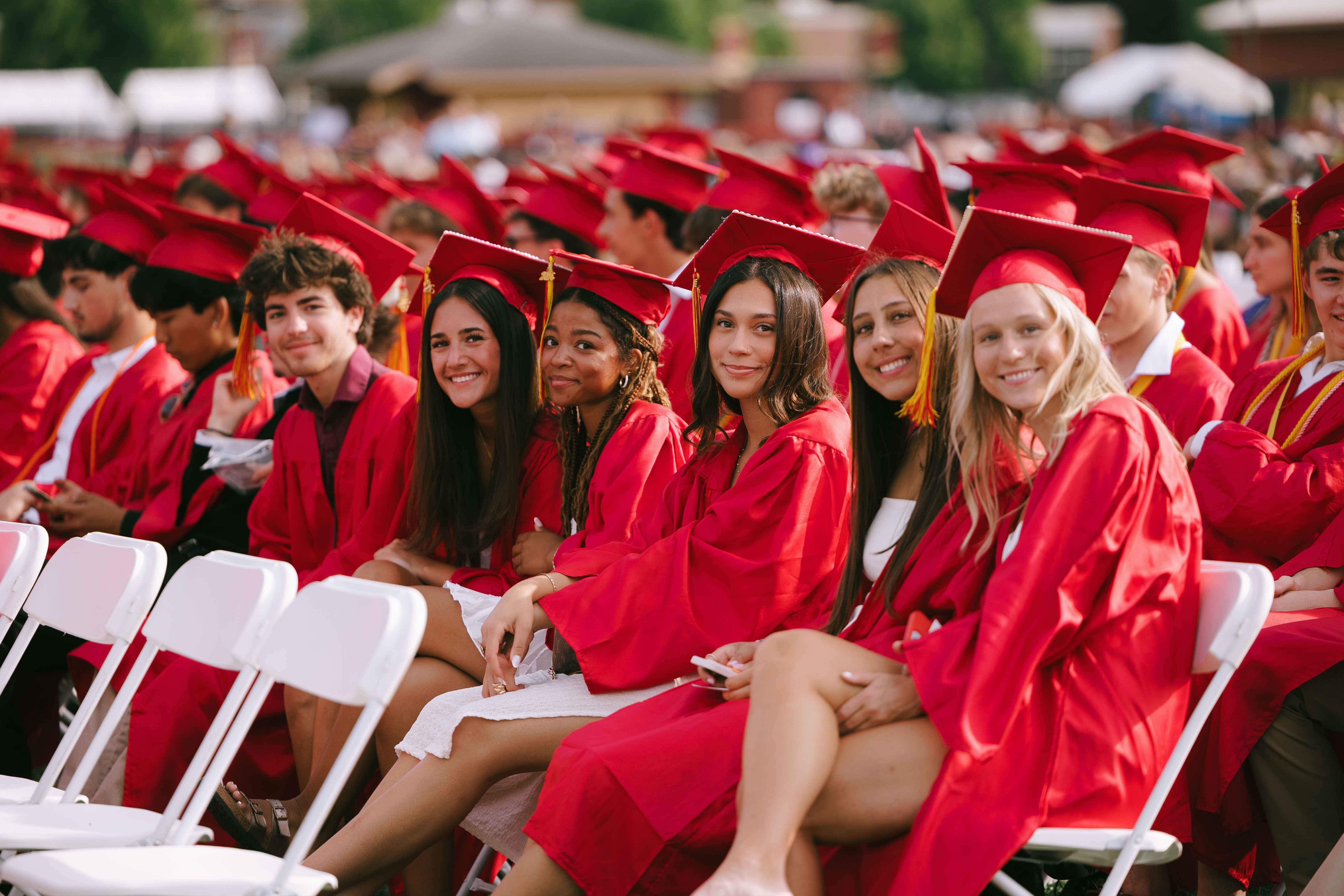 Several students in red graduation gowns and caps sitting on chairs in a large outdoor venue.