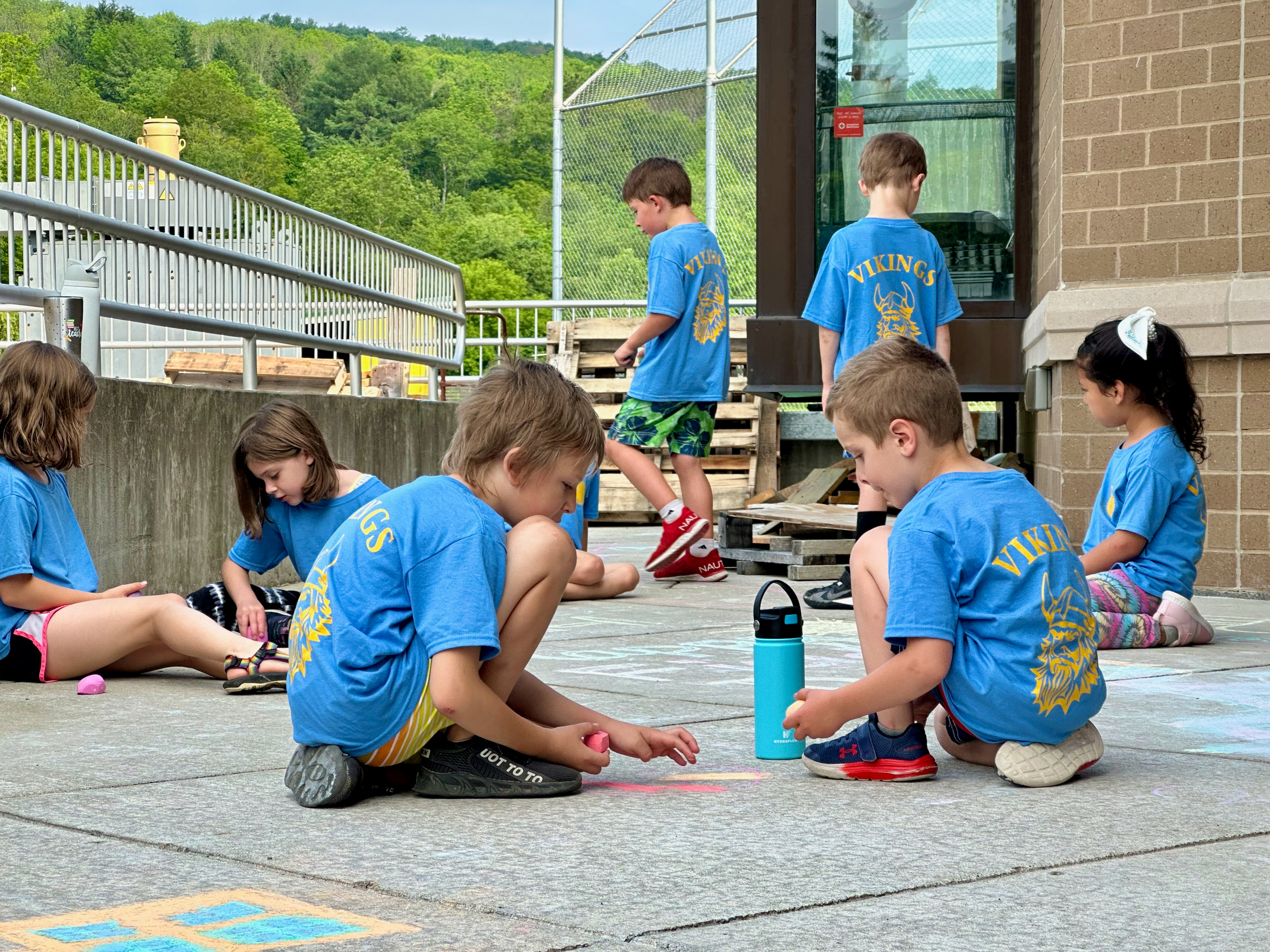 Elementary students in matching blue shirts make chalk drawings  outside school during Field Day