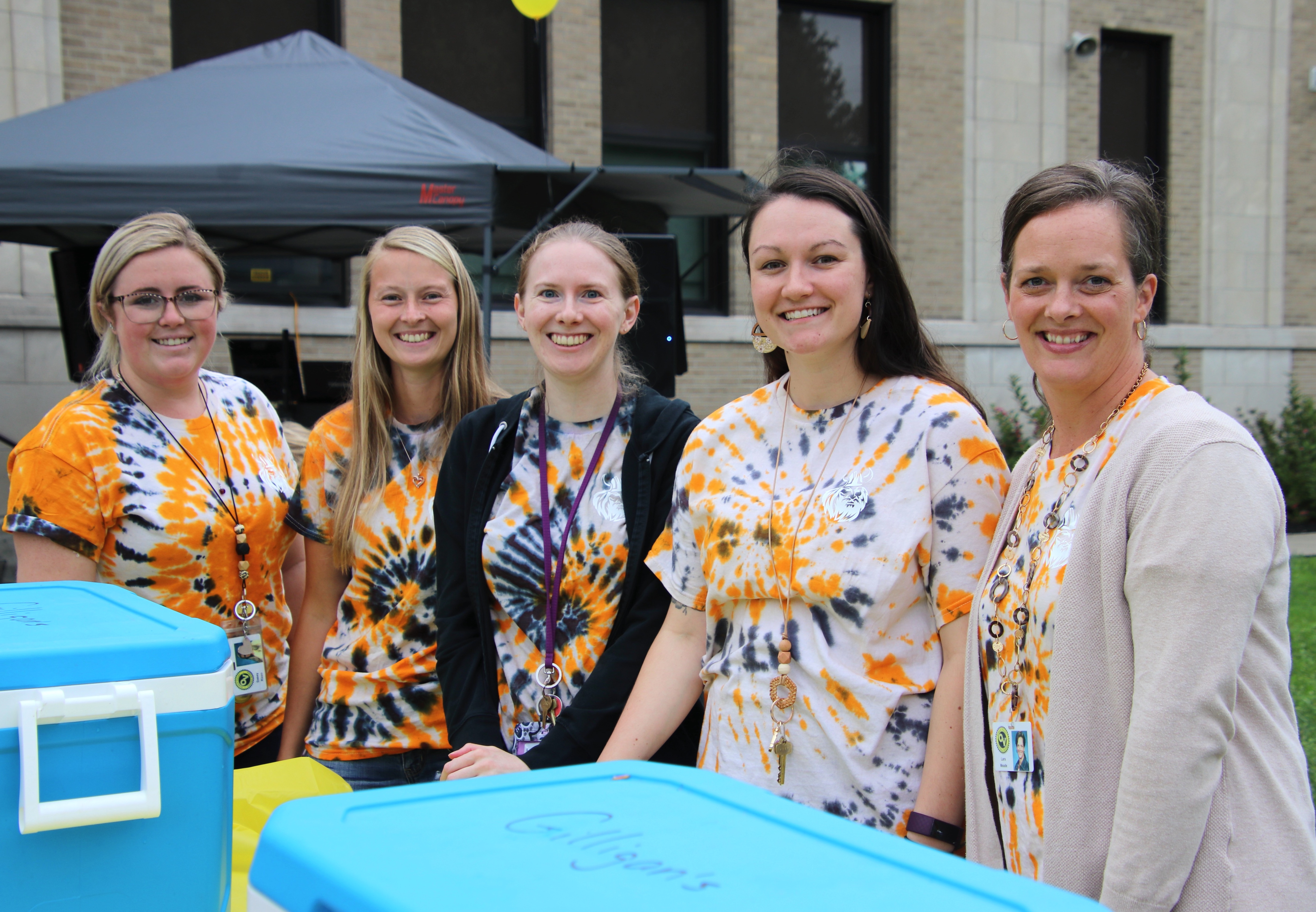 A group of 5 young female teachers all wearing tie-dye t-shirts stand outside a school building at a special back-to-school event