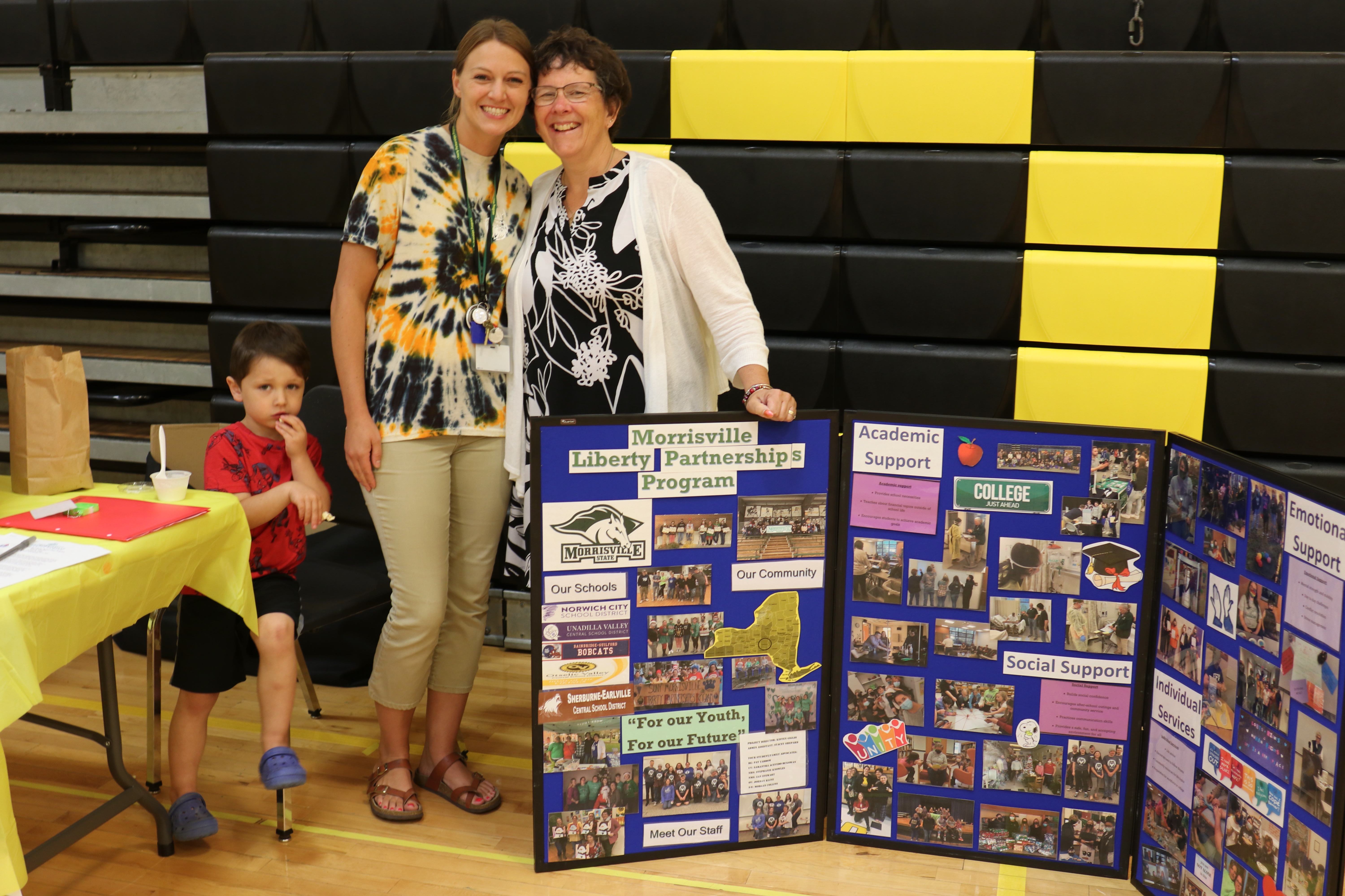 Two female educators stand in a school gym with a big poster featuring  Liberty Partnership, a school program providing extra help to students