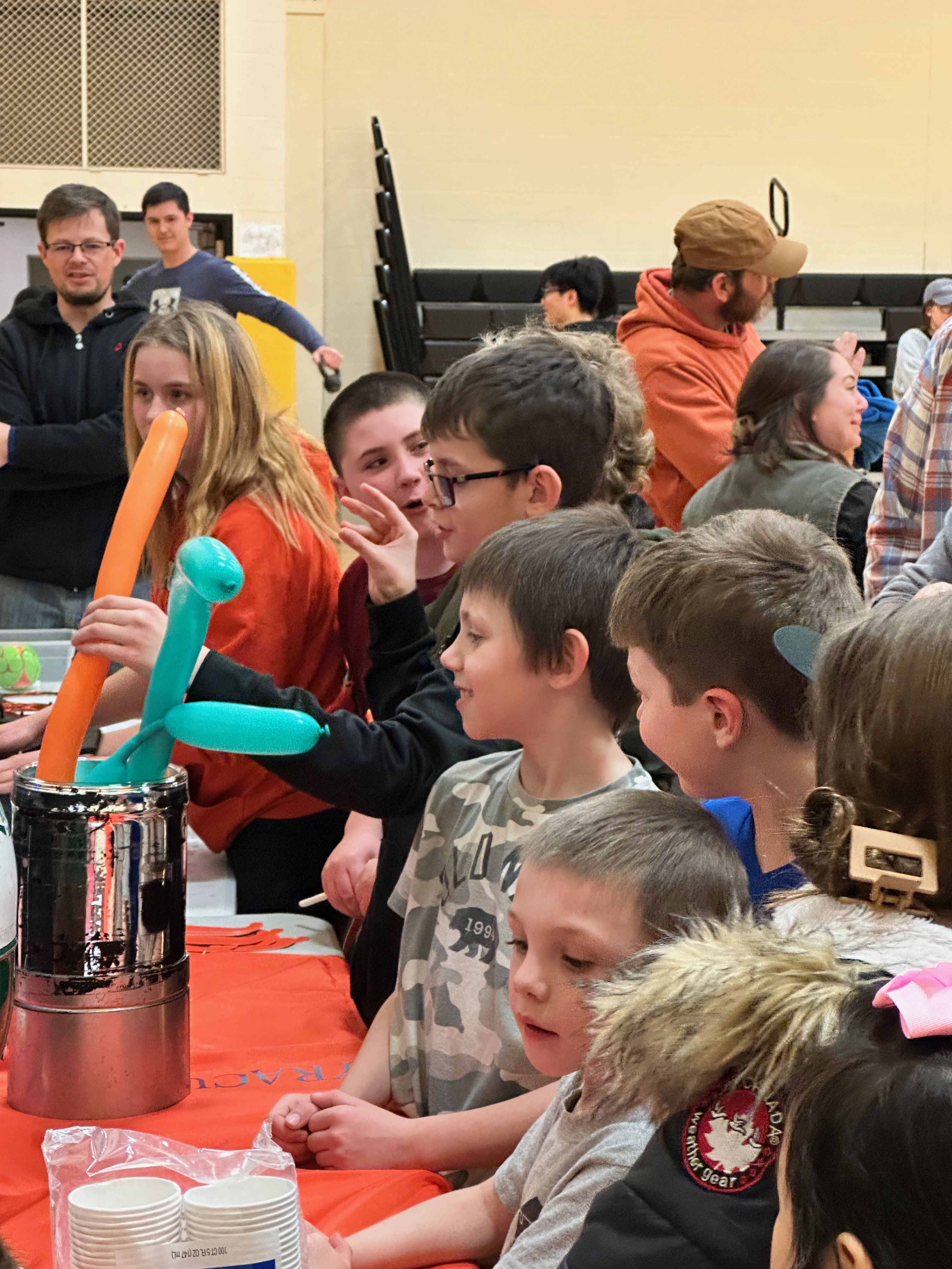 A bunch of students cluster at a display table at a Science Night event, where a college student is working with balloons to demonstrate chemistry and physics