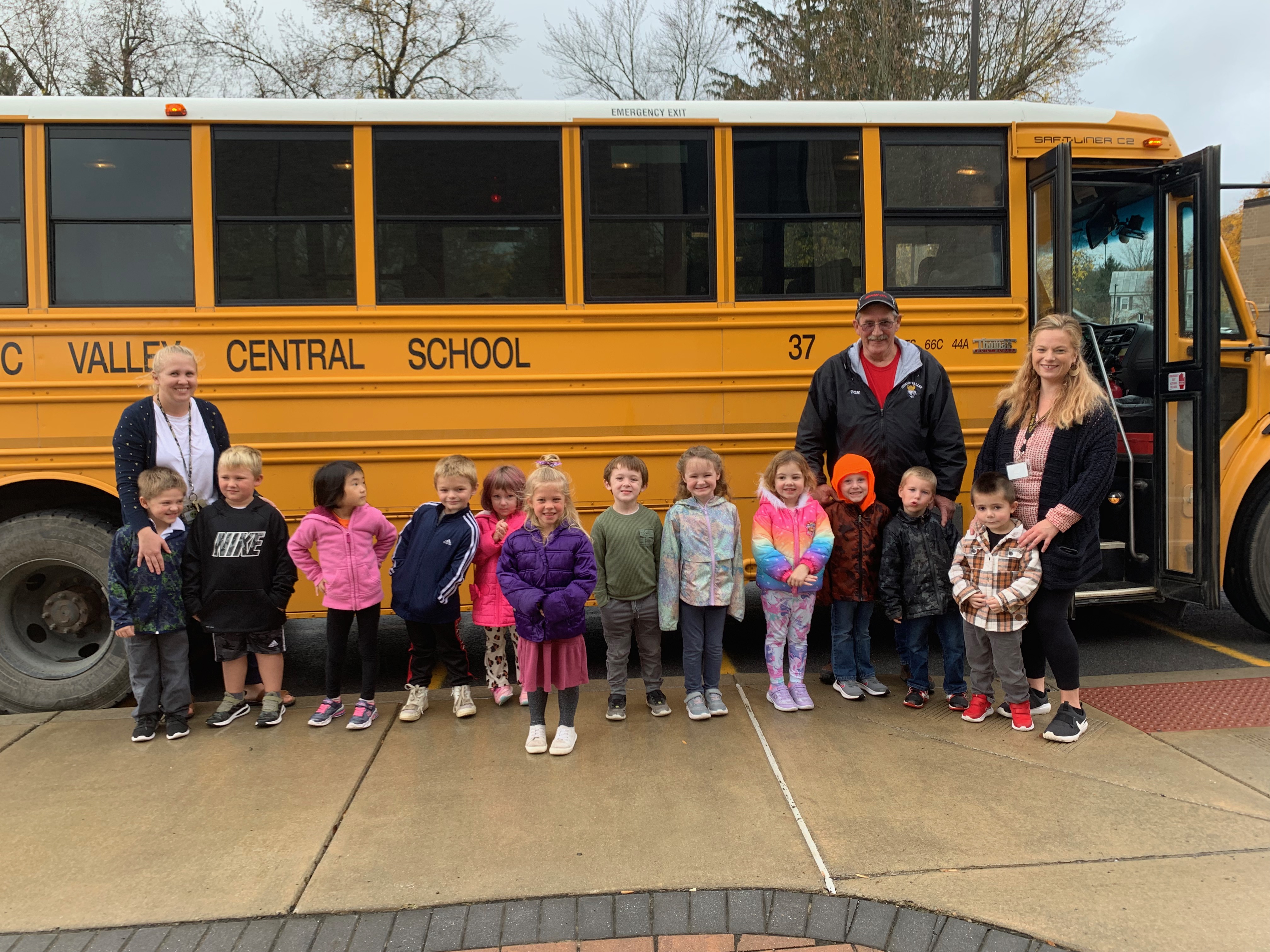 A preschool class in fall jackets stand in front of a school with their teachers and the bus driver after their bus safety tour of the bus