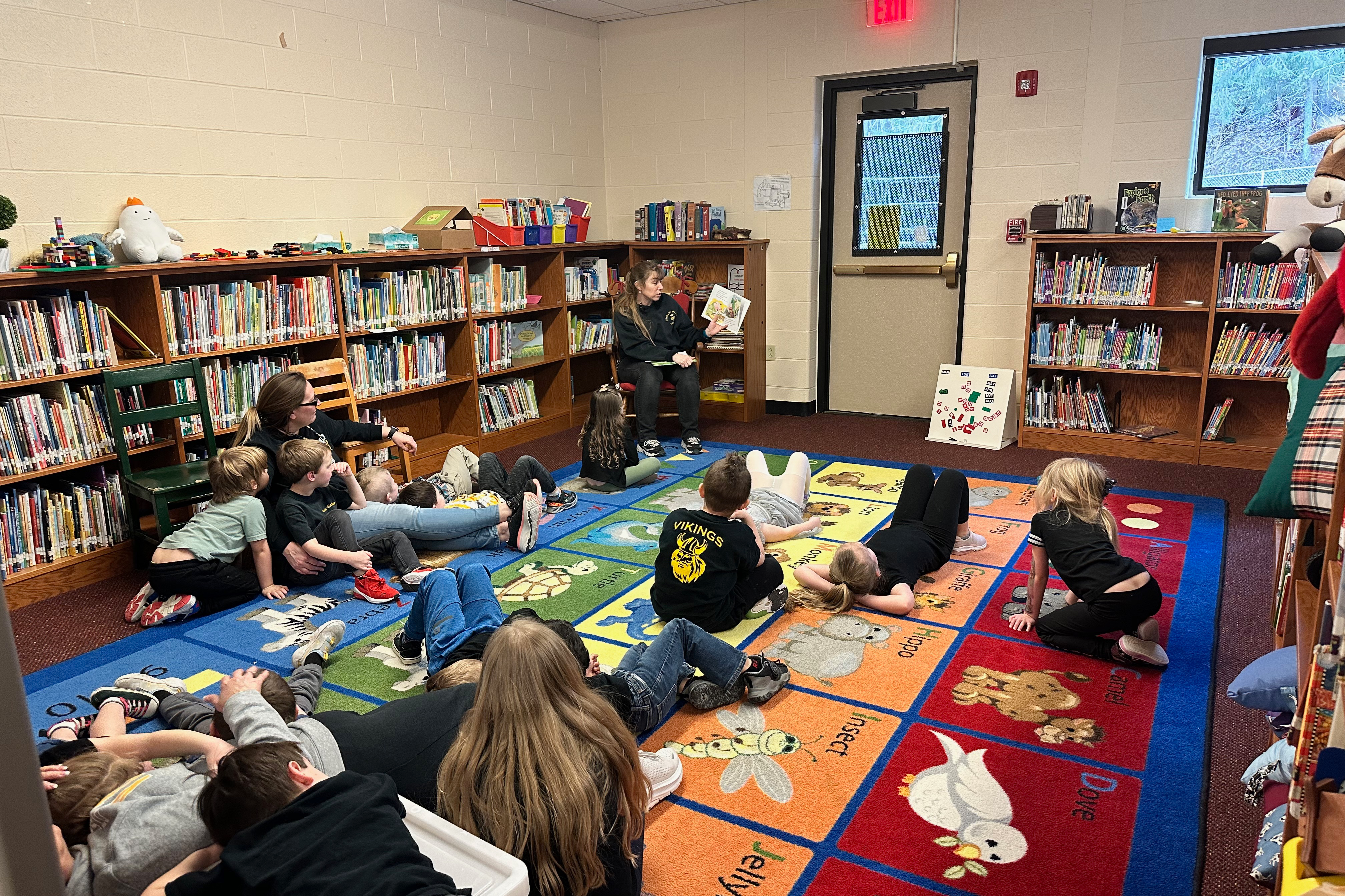 Young students sit and lay down on a colorful library rug while an aide reads to them as she sits in a rocking chair 