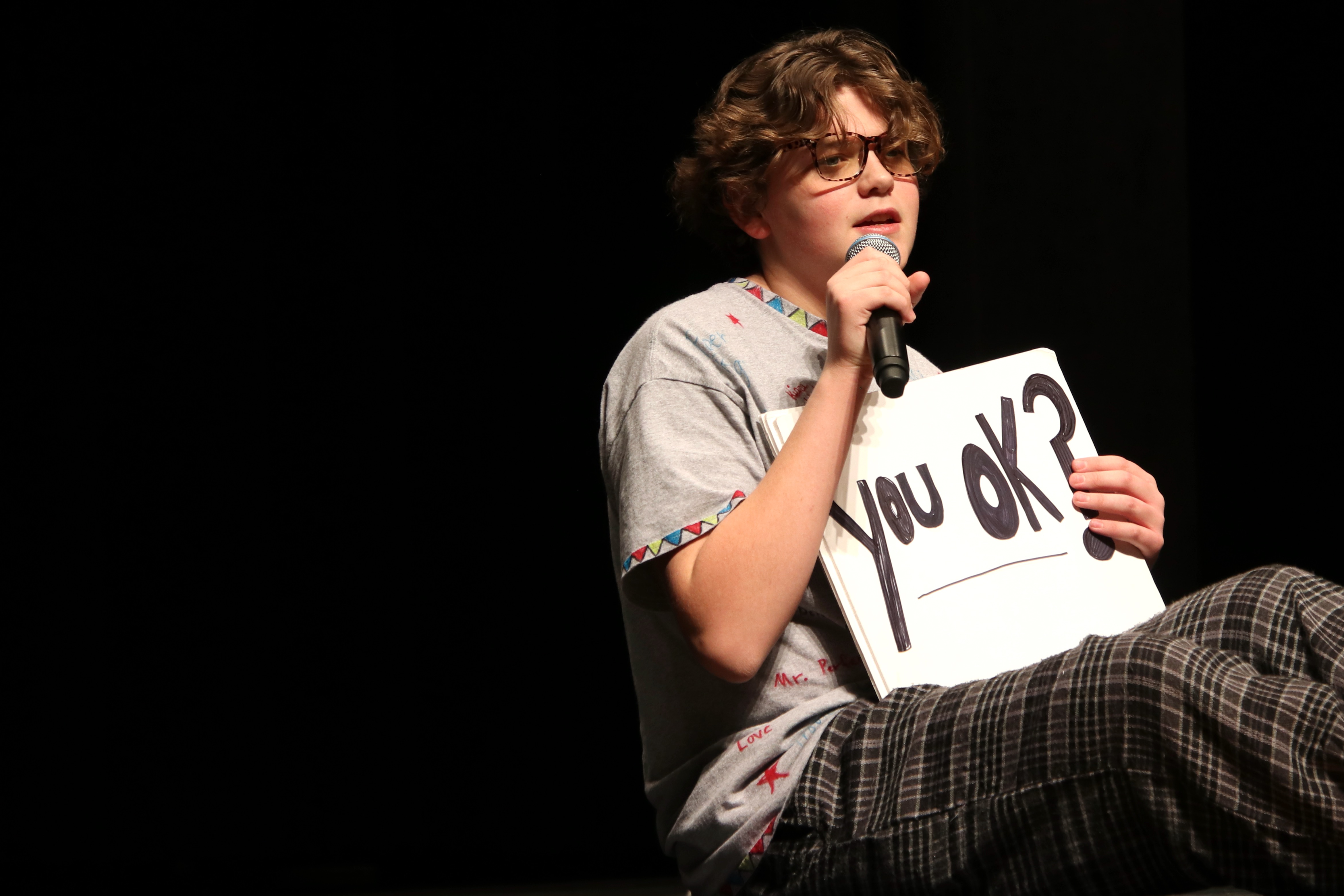 A teenage boy on a school stage with a microphone holds a sign that says "You okay?"