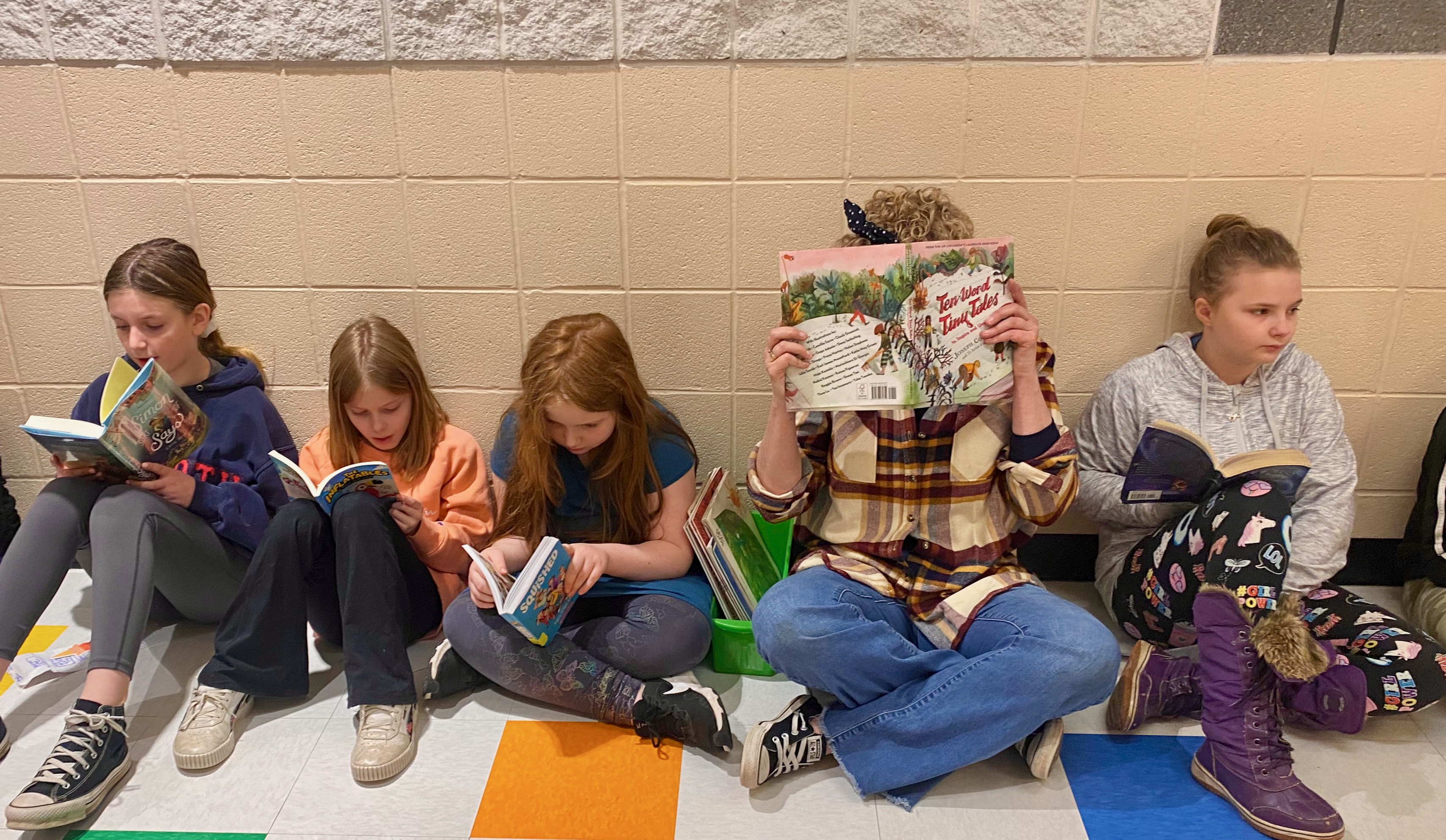 Students and a teacher sit on the floor in a classroom hallway while reading books