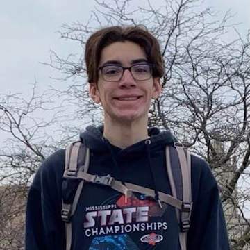 Photo of Mateo Byrd in front of trees. He is wearing a backpack and wearing glasses and a swim championship hoodie. 