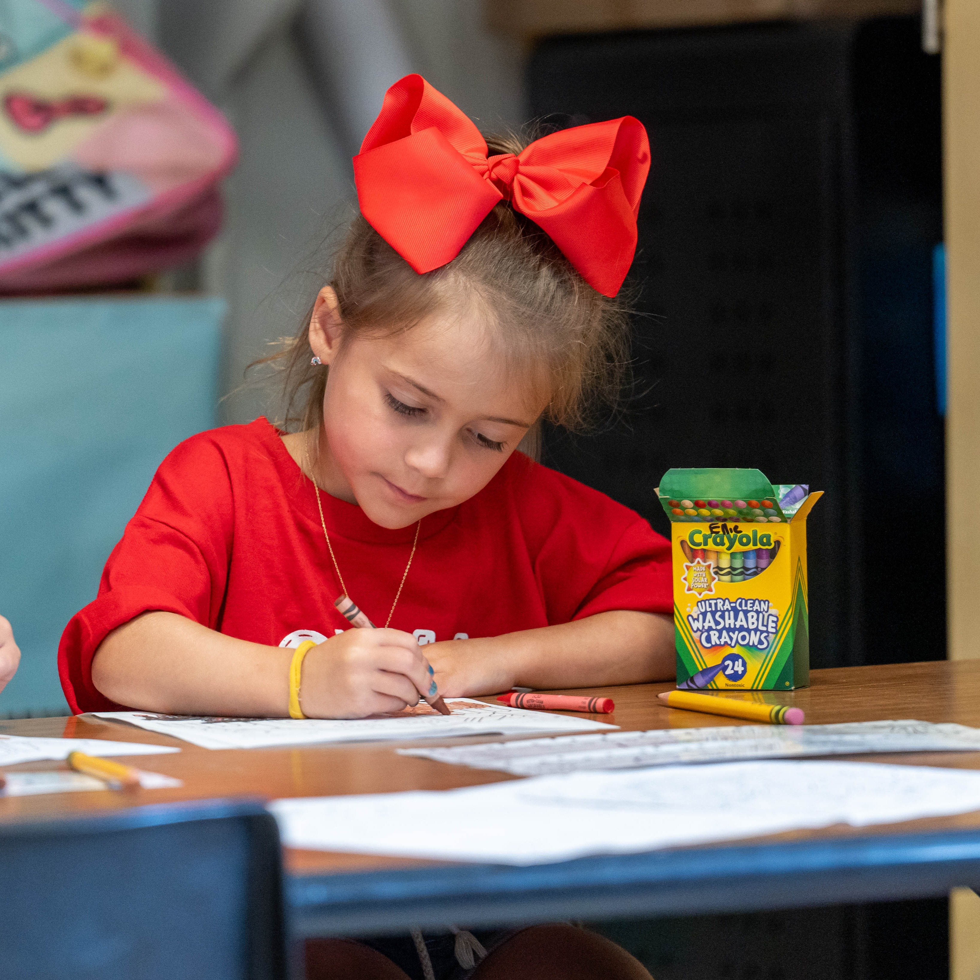 A young child in a red shirt sits at a table, coloring with crayons and papers.