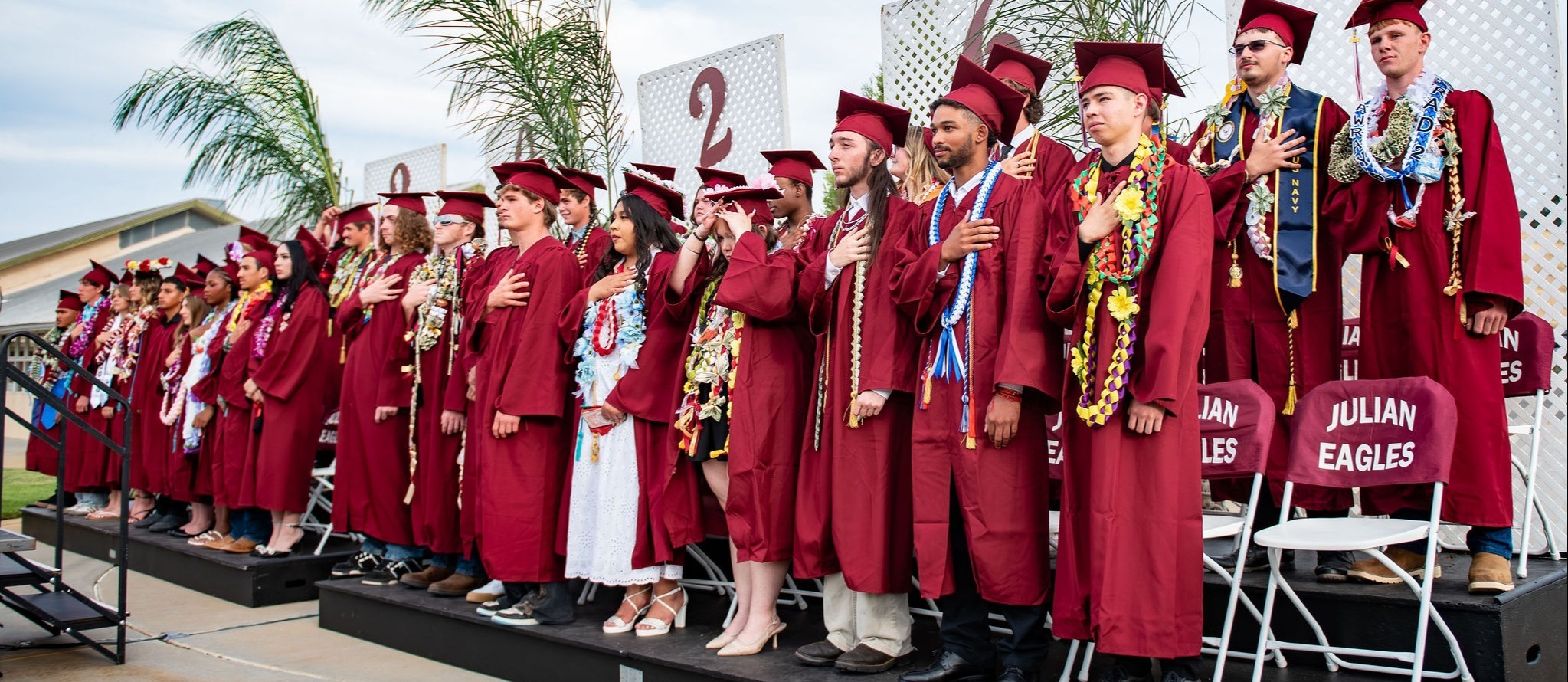Graduates in red robes and caps stand on a stage in front of a building with palm trees.