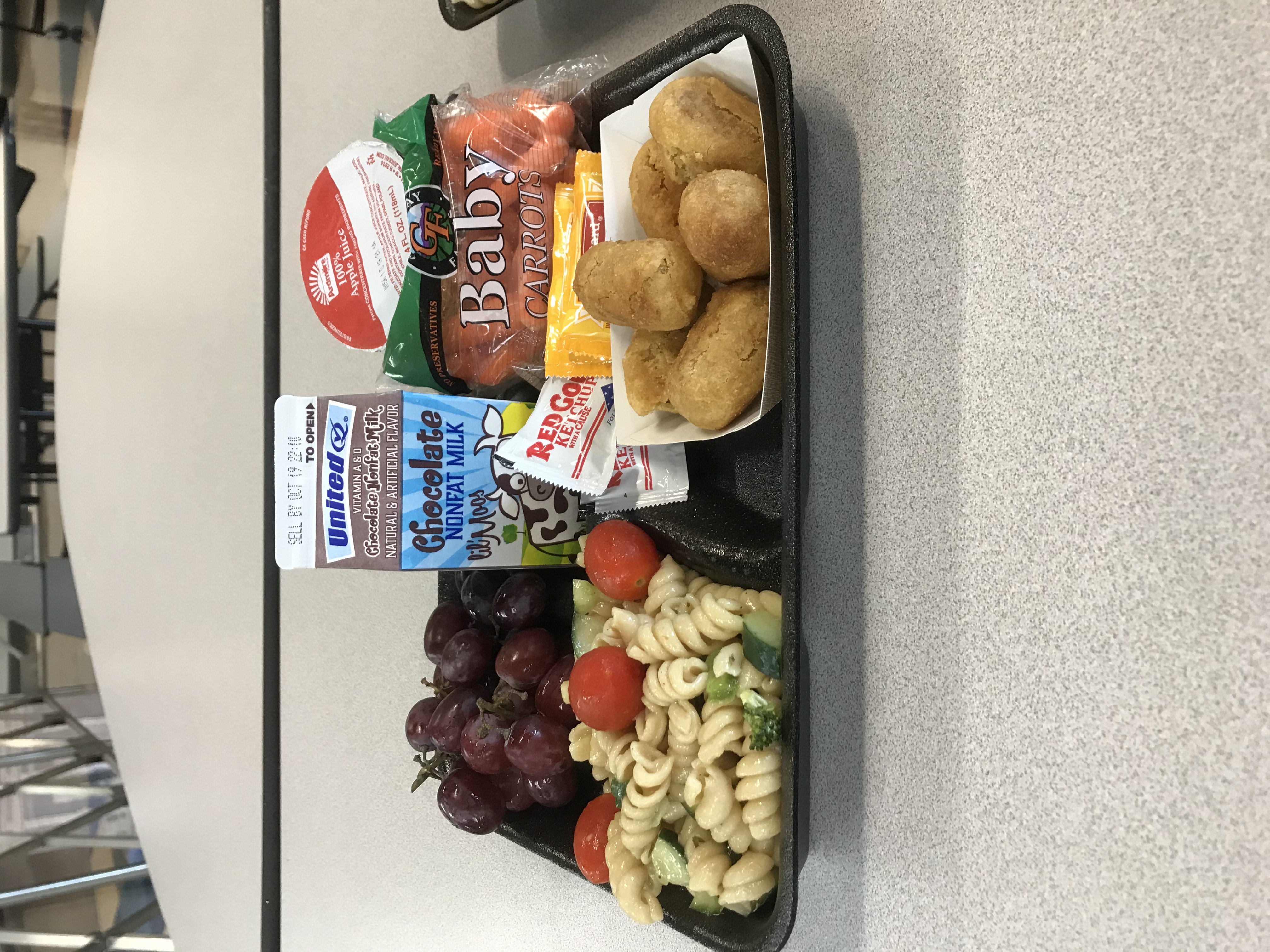 A tray of food containing mini corn dogs, pasta salad, grapes, a carton of milk, baby carrots, and yogurt.