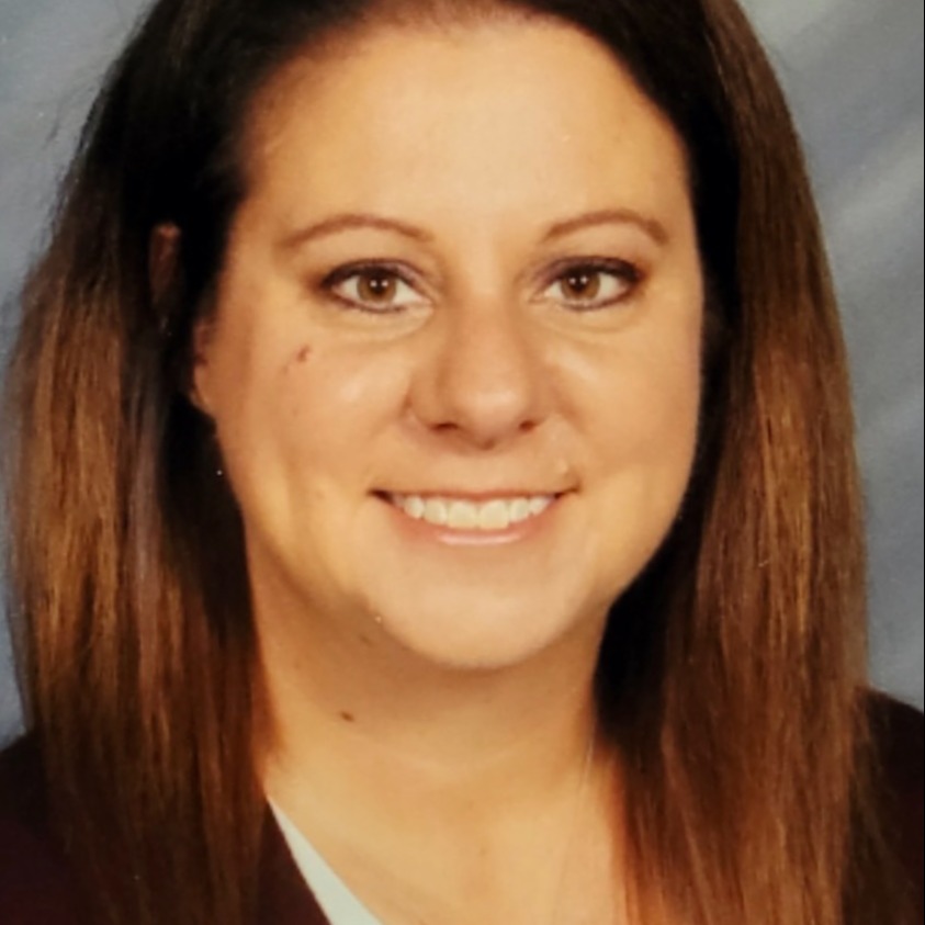 A woman with long, brown hair and a white shirt, smiling and showing teeth against a plain background.