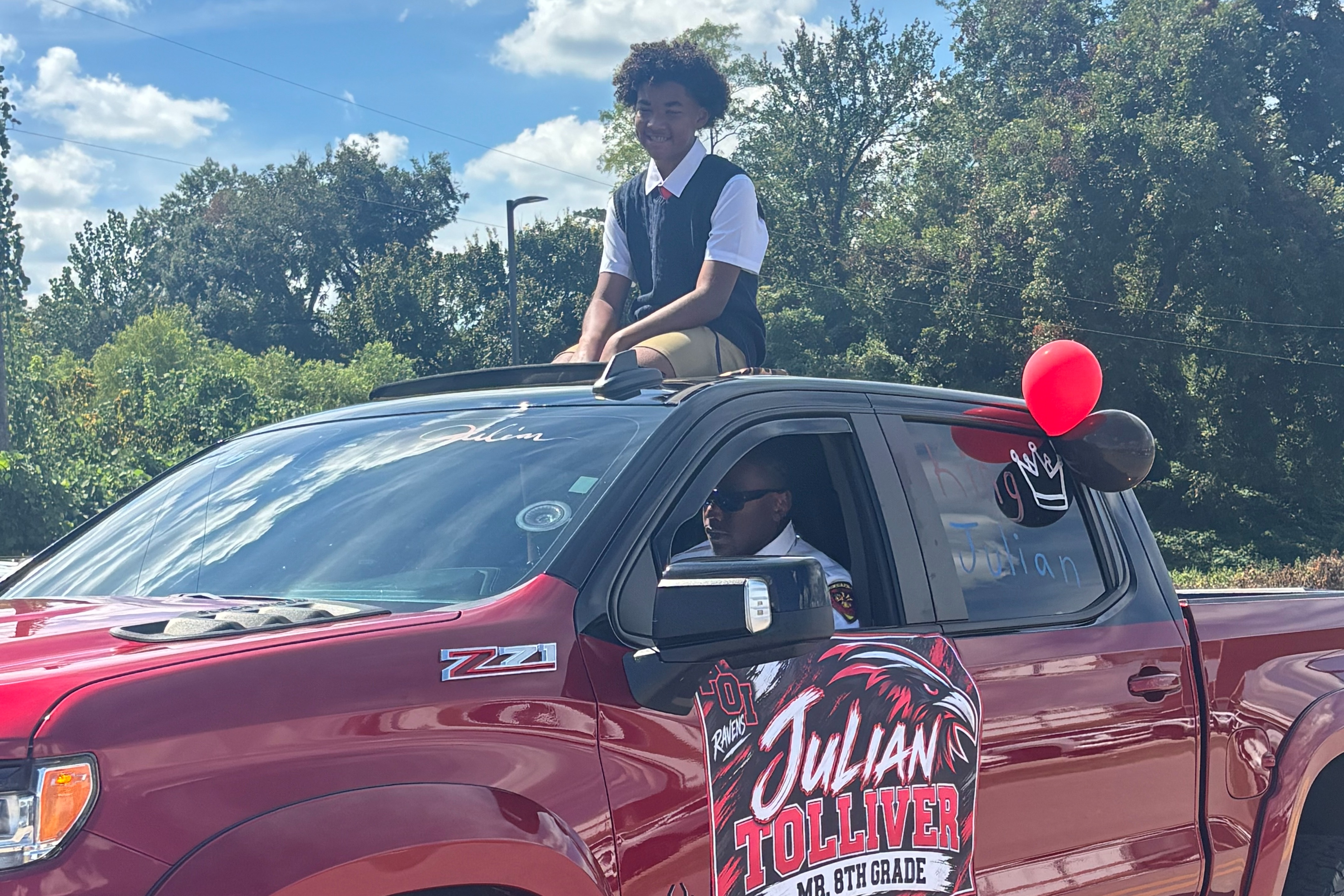Red pickup truck with a person sitting inside and another on the hood. A banner on the truck reads "Julian Talento."