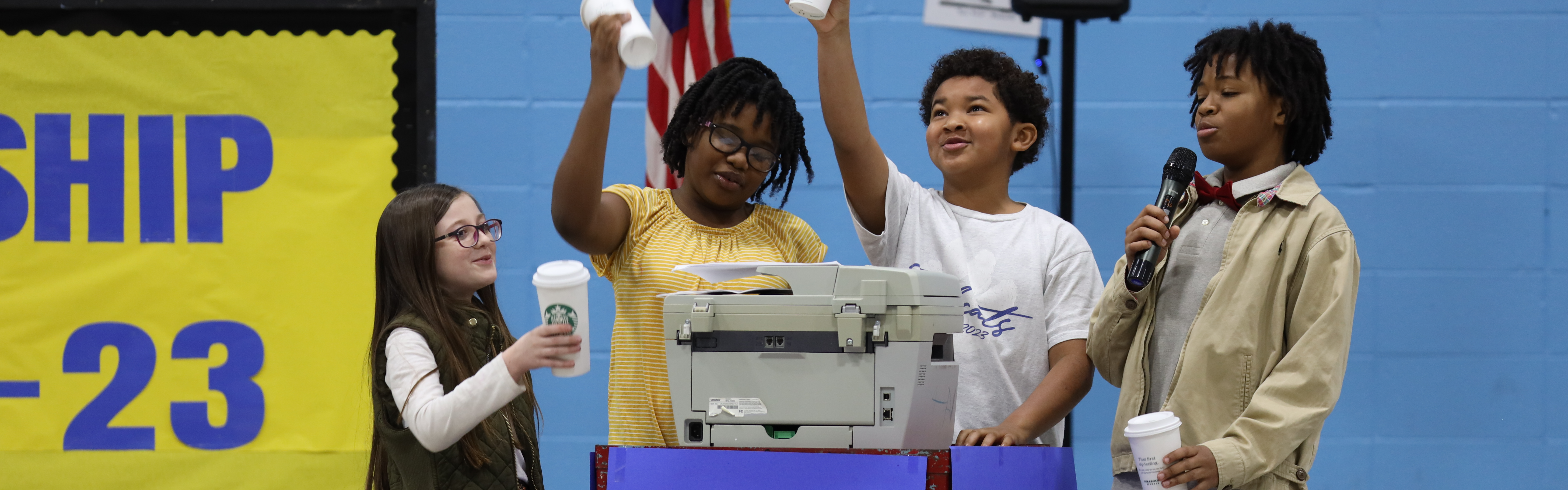 Four students are performing a skit. Students are acting as teachers and are raising coffee cups up in celebration. The students are standing behind a cart.